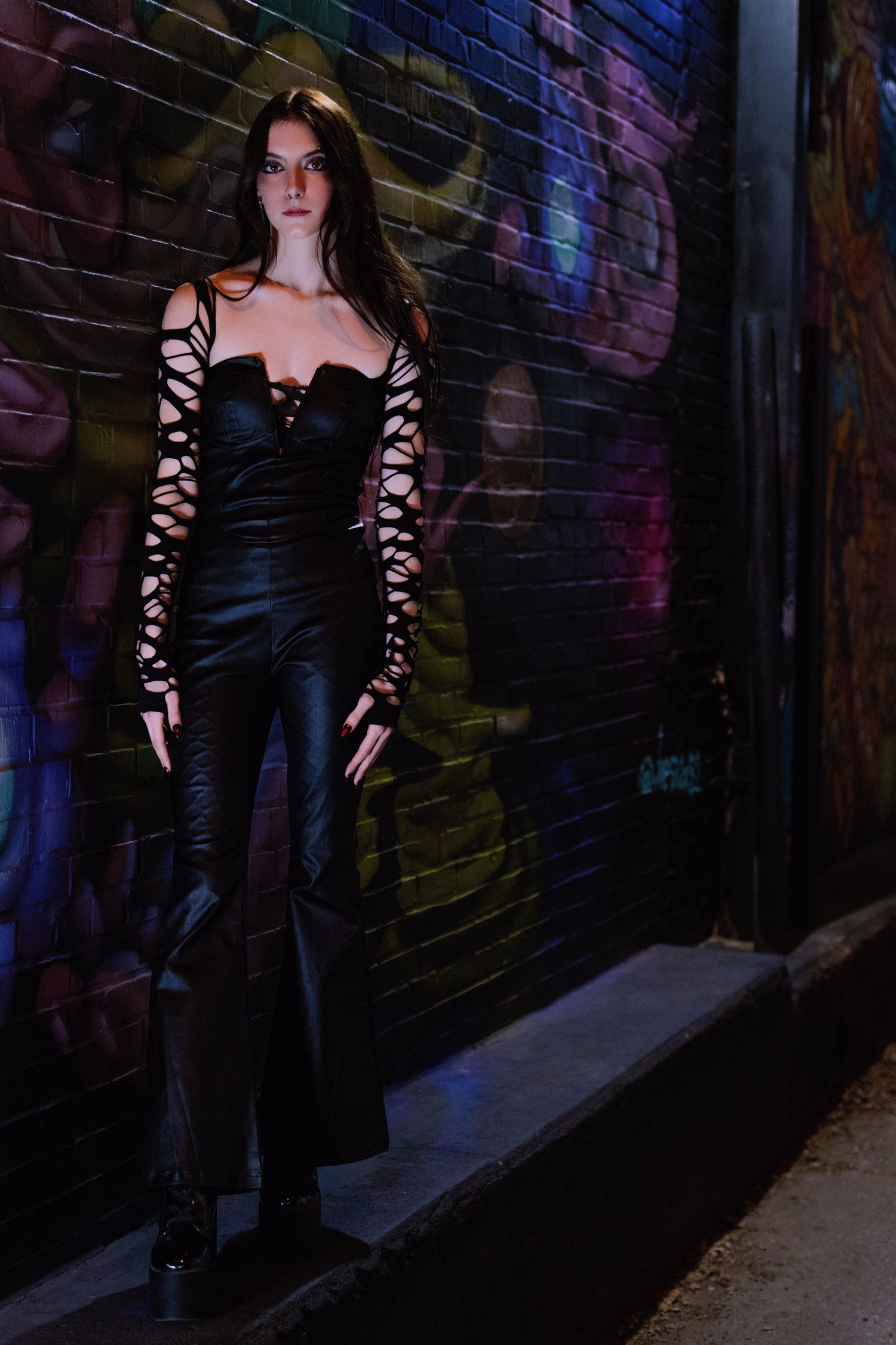 Woman in black lace-sleeve corset jumpsuit stands against graffiti brick wall at night.