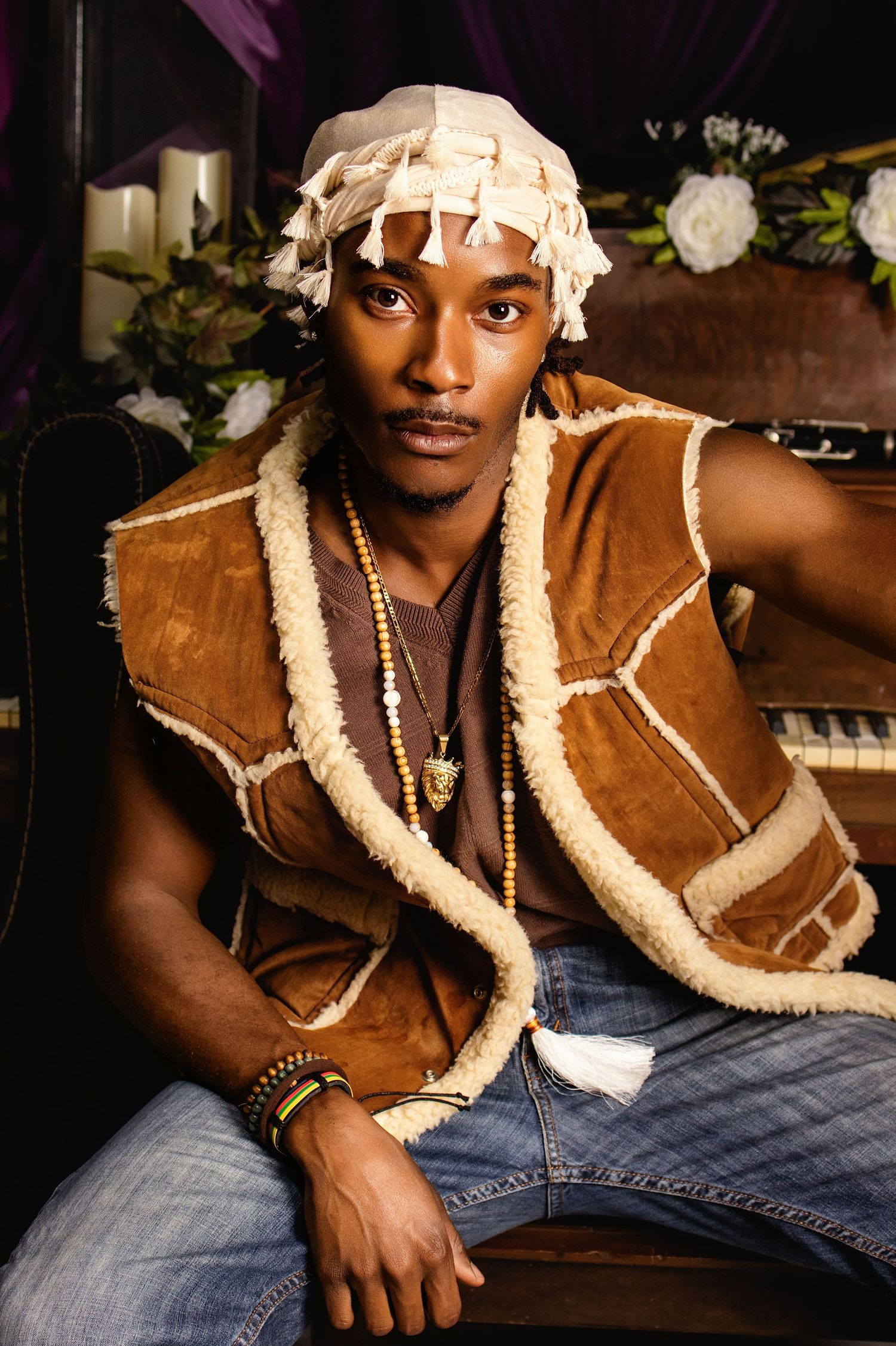 Young Black man in shearling vest and white head wrap poses near vintage piano with purple drapes.