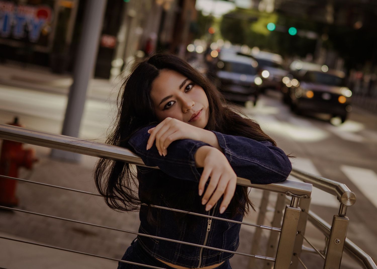 Young woman with dark hair leans on metal railing on city sidewalk at dusk with street traffic behind.