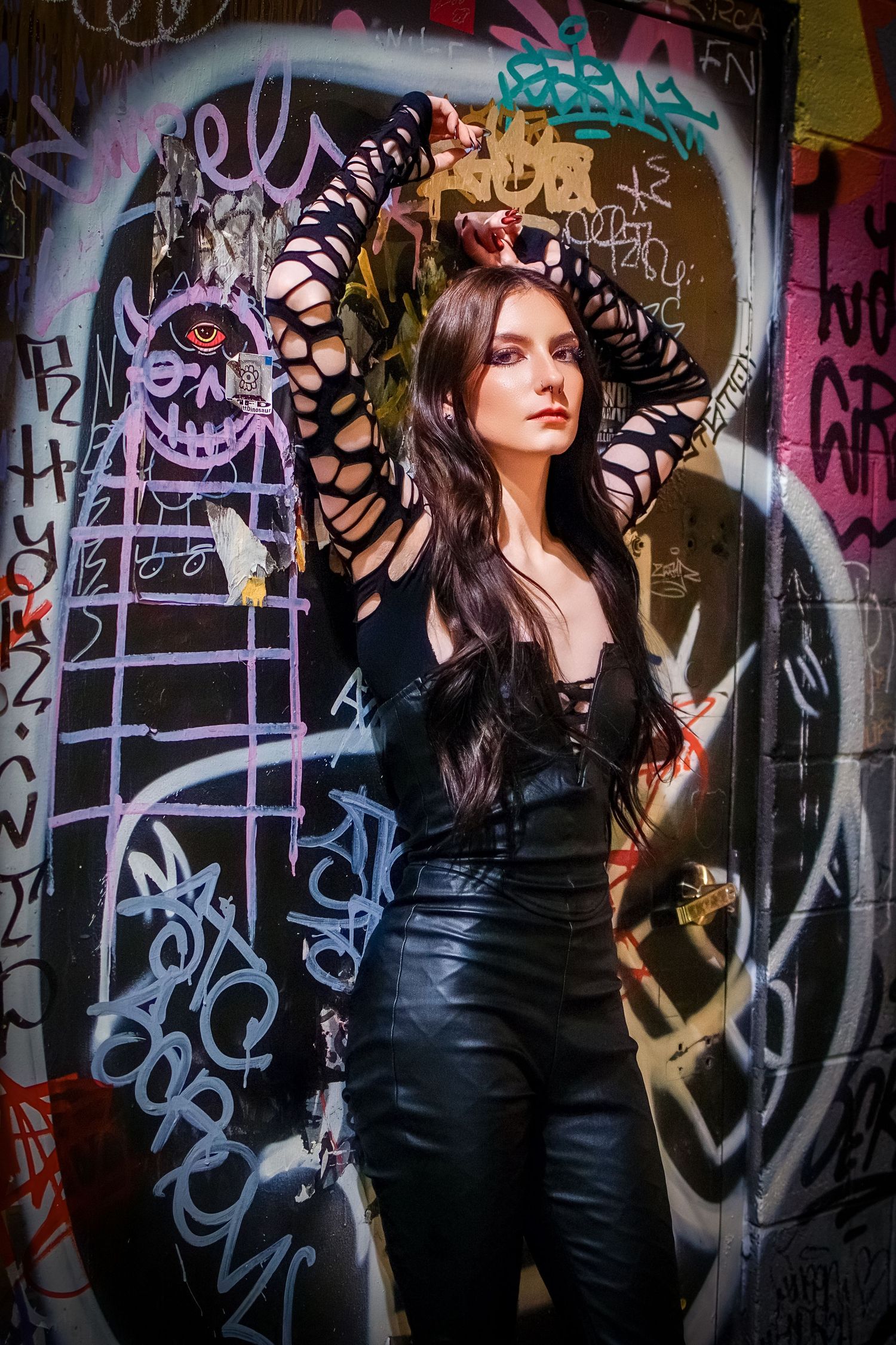 Woman in black cutout sleeves and leather pants posing against colorful graffiti wall.