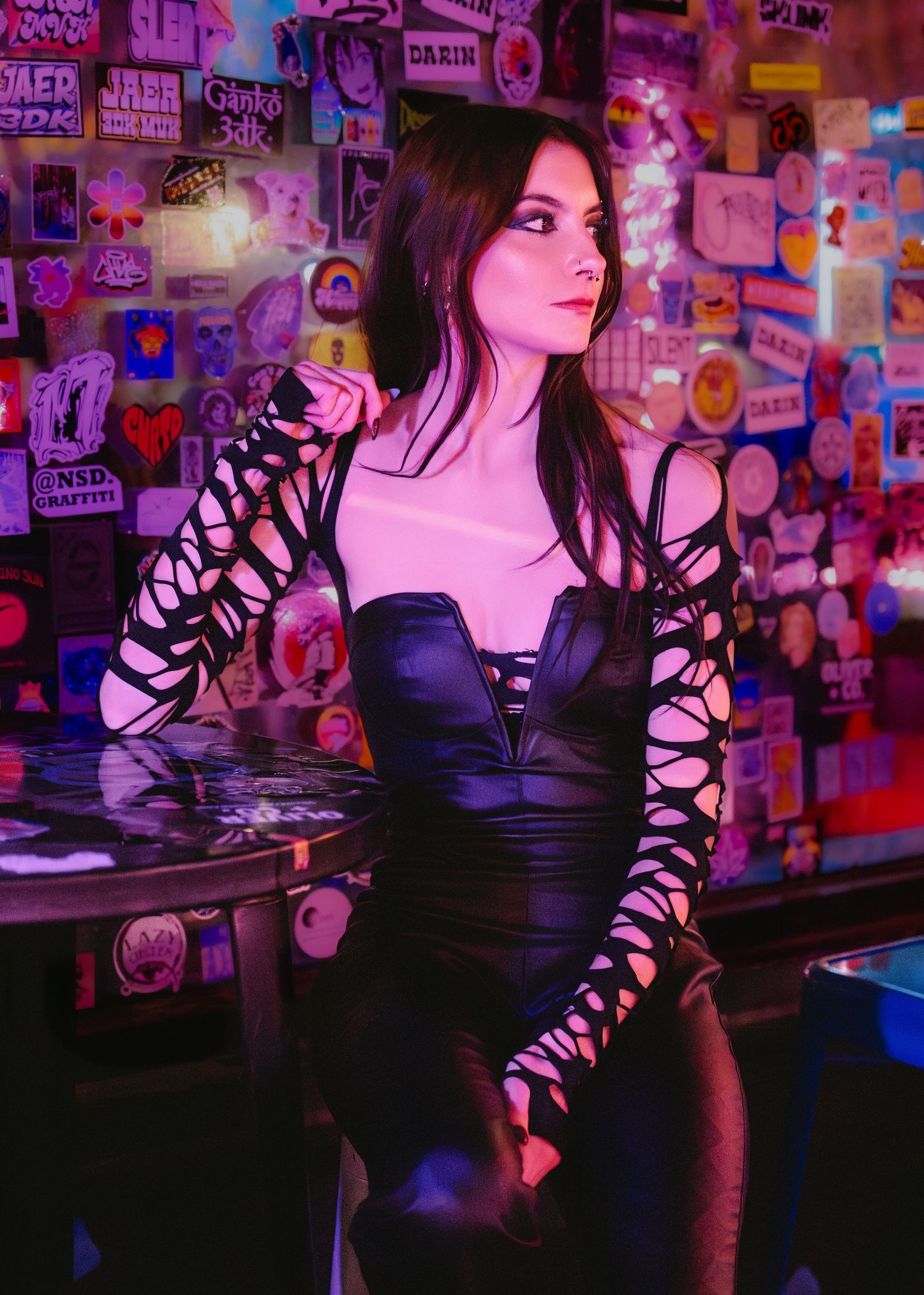 Woman in black lace cutout dress poses against colorful sticker wall under neon pink lighting.