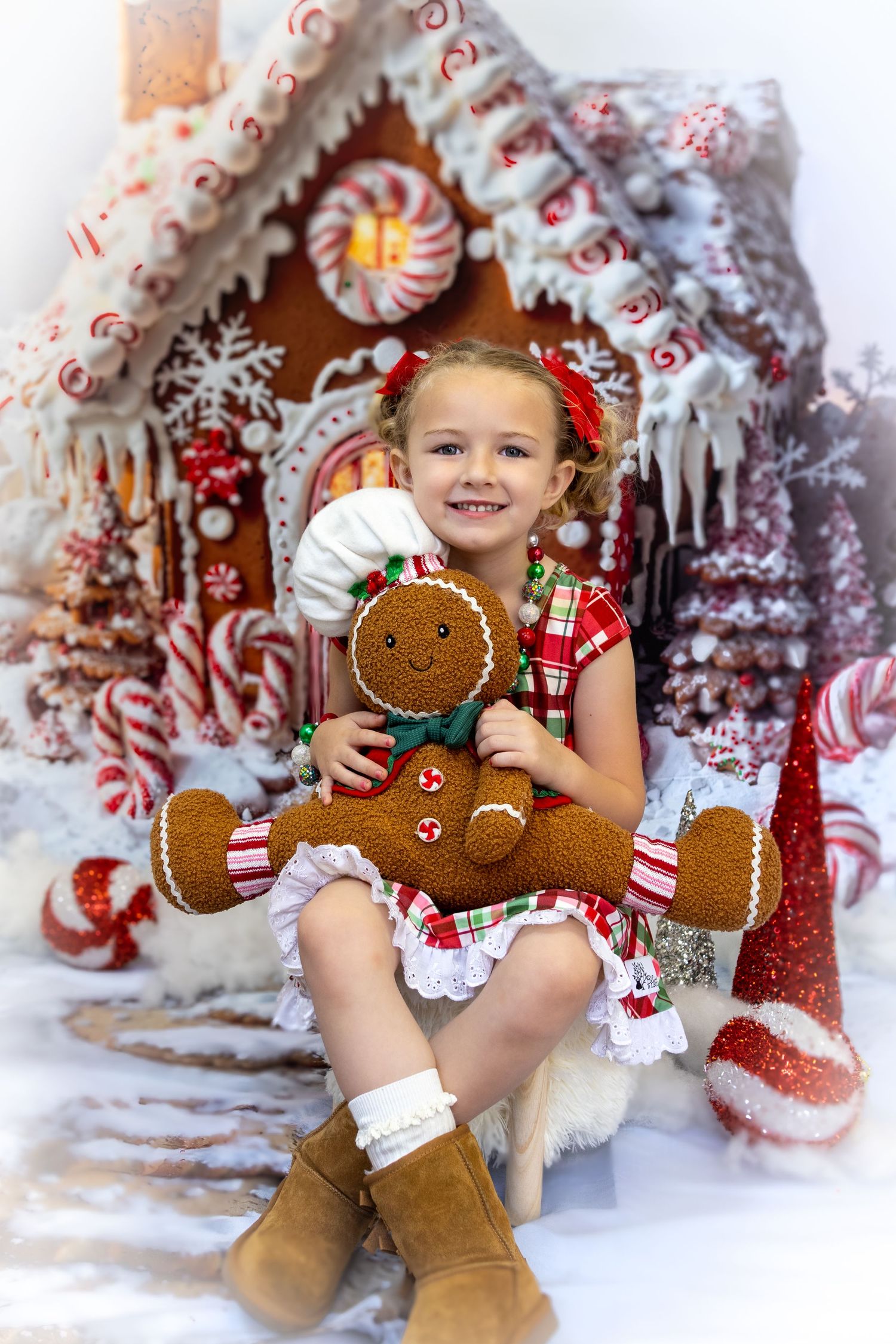 A smiling girl in a festive dress holds a gingerbread man plush toy in front of a decorated candy gingerbread house.