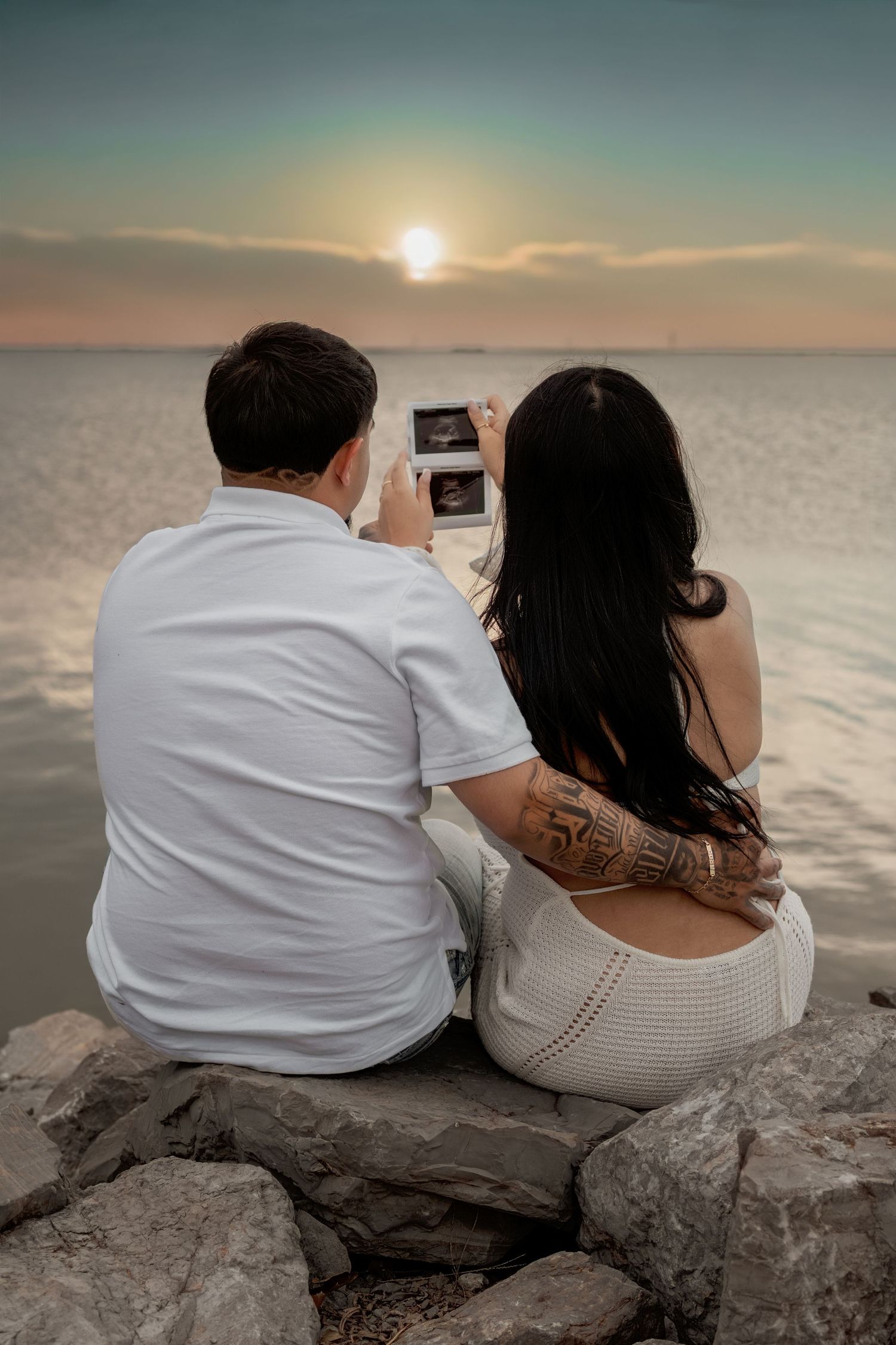 Couple sitting on rocks by the ocean at sunset, holding an ultrasound photo, announcing their pregnancy.