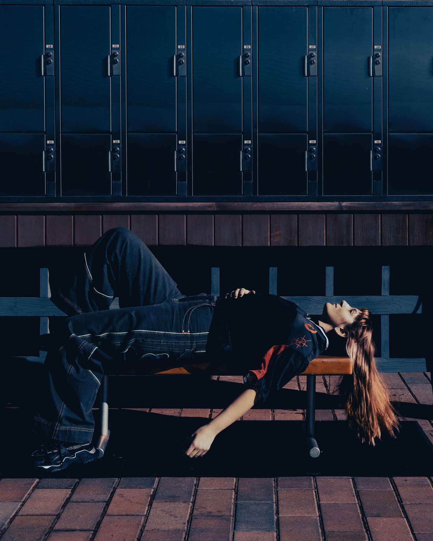 Person lying on a bench in front of blue school lockers, wearing jeans and a red jacket.