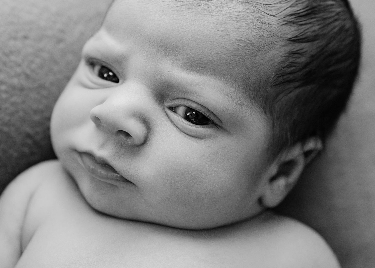 Black and white close-up portrait of a newborn baby with dark hair, looking upward with wide eyes.
