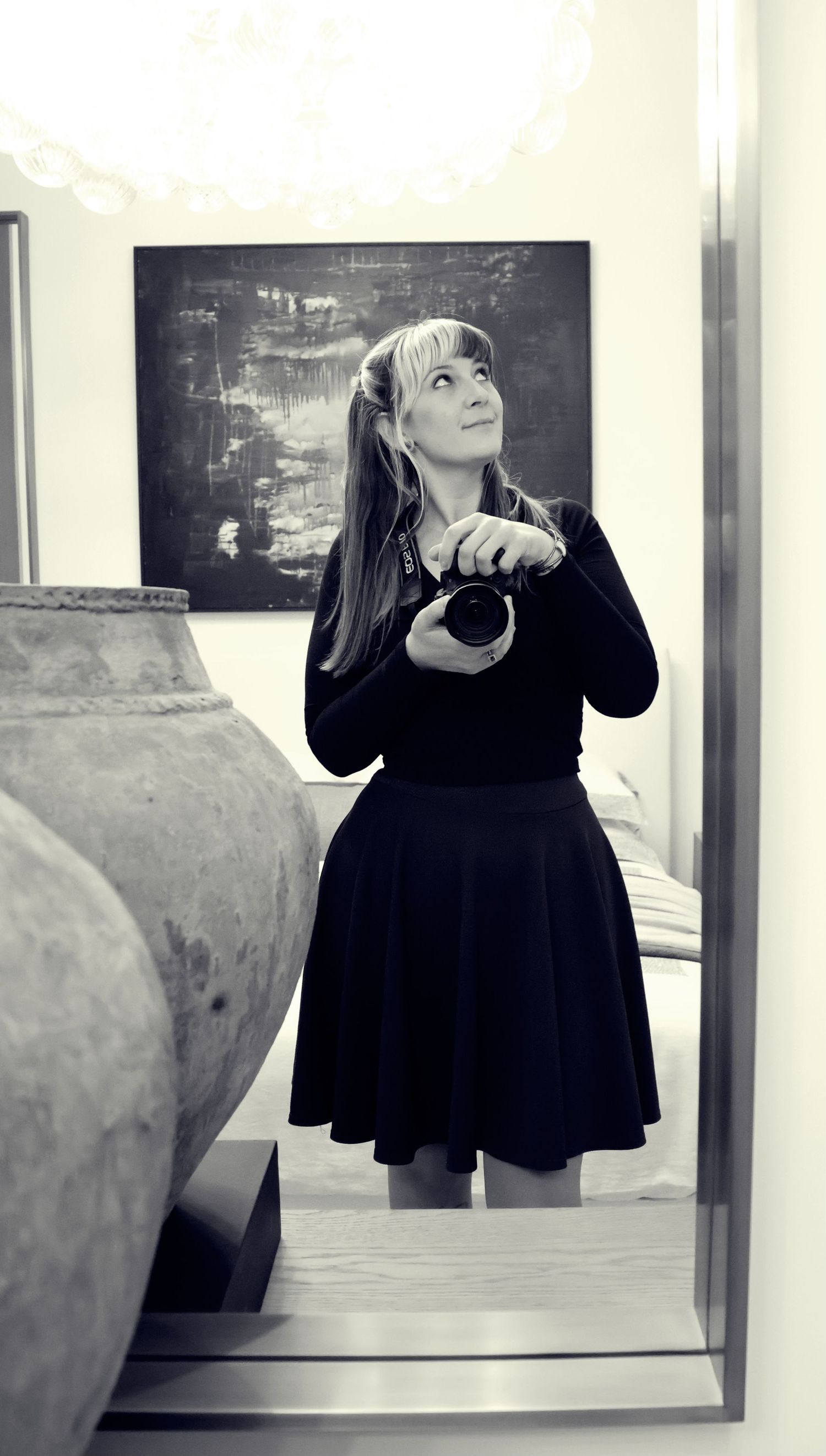Black and white photo of a woman in a black dress holding a camera in a museum with ancient pottery and abstract art.
