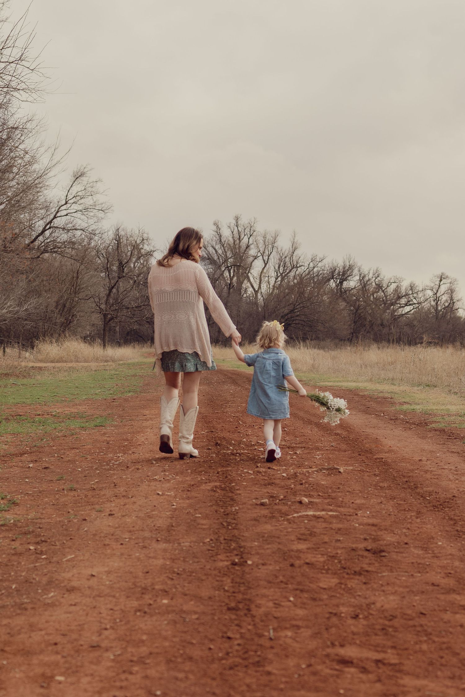 Mother and young daughter holding hands walking down a rustic dirt path surrounded by bare trees.