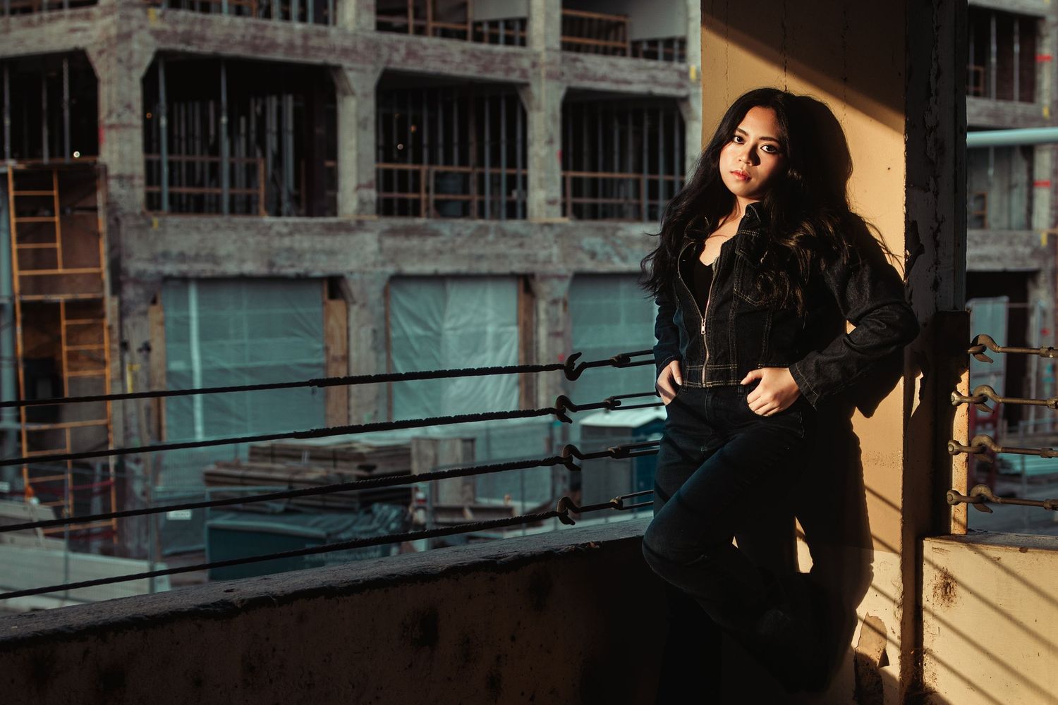 Young woman in all-black outfit posing on rooftop with industrial building backdrop.