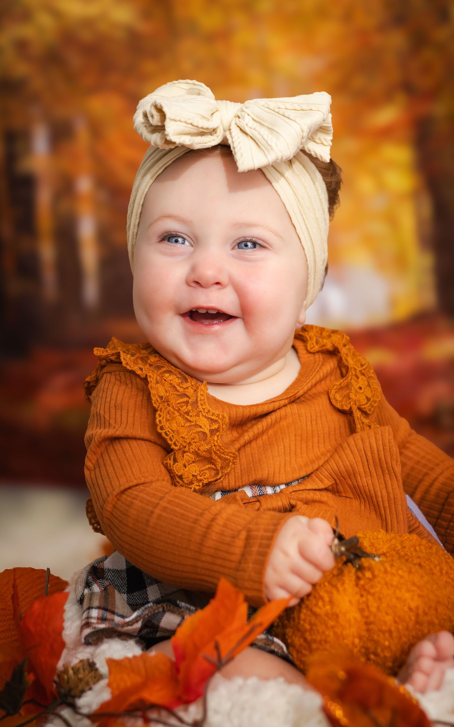 Smiling baby in rust orange outfit with cream bow headband, surrounded by fall leaves and pumpkins.