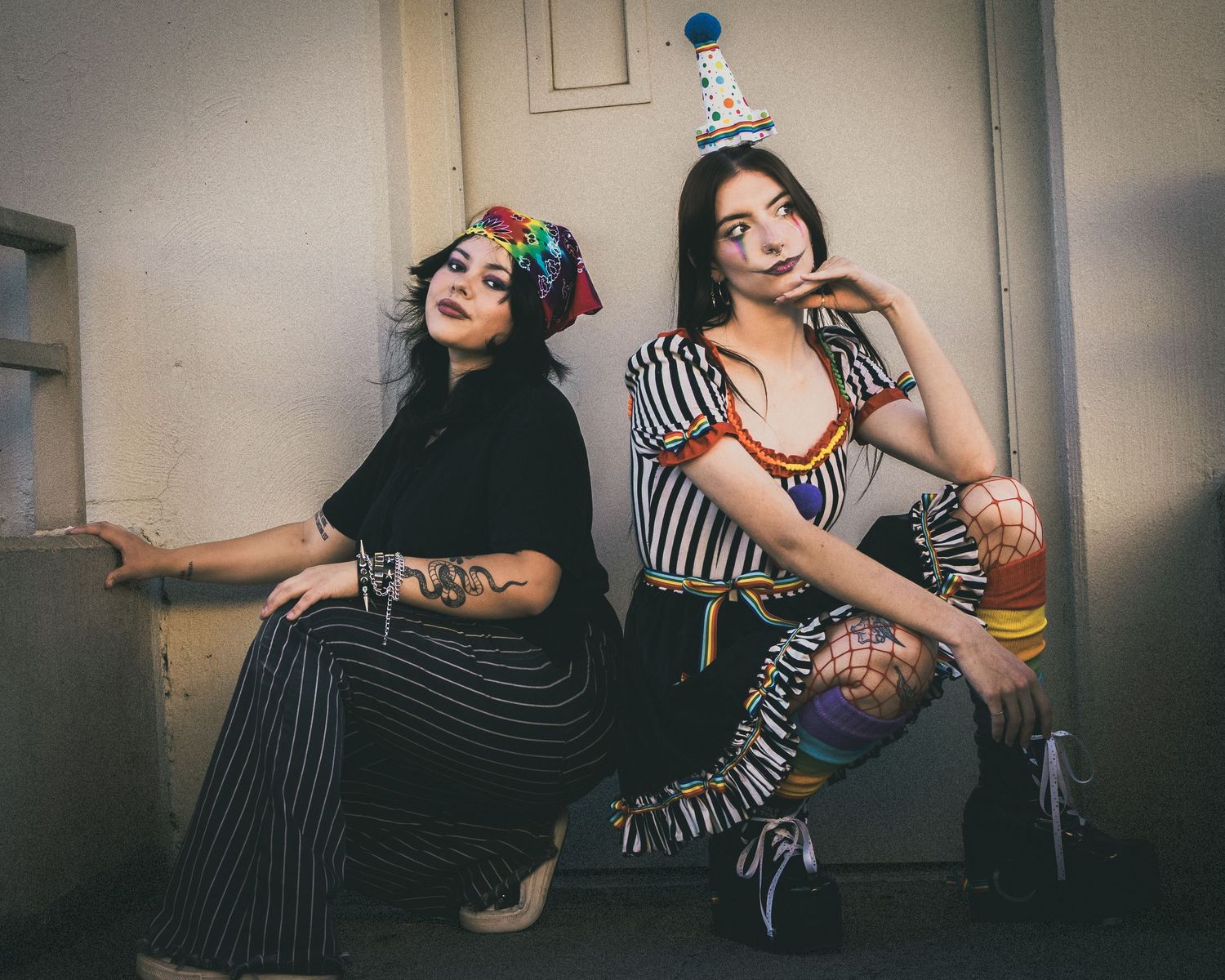 Two women in clown-inspired costumes with face paint and party hats pose dramatically against a white wall.
