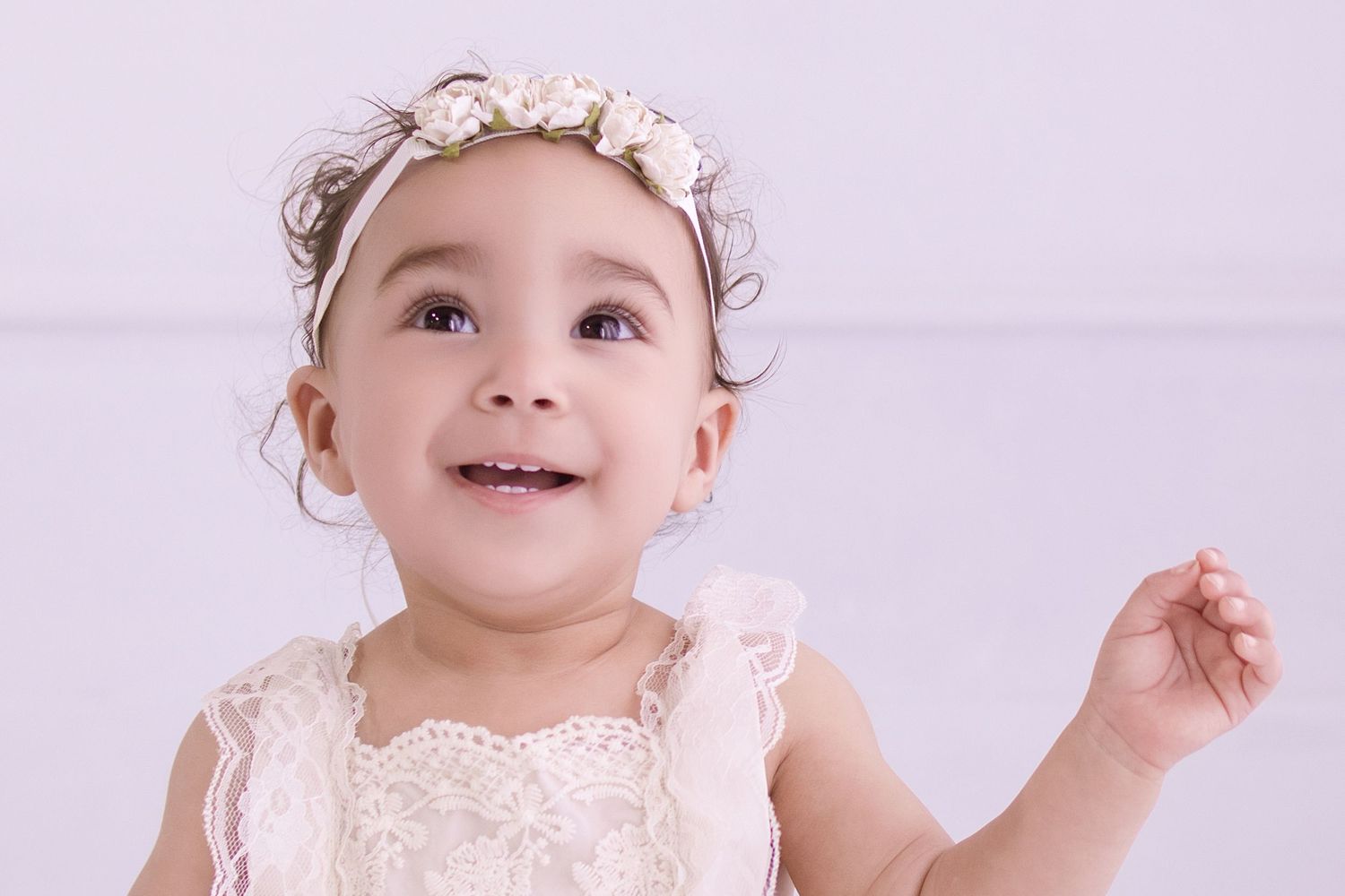 Adorable baby girl in white lace dress and floral headband smiling and looking up against light background.