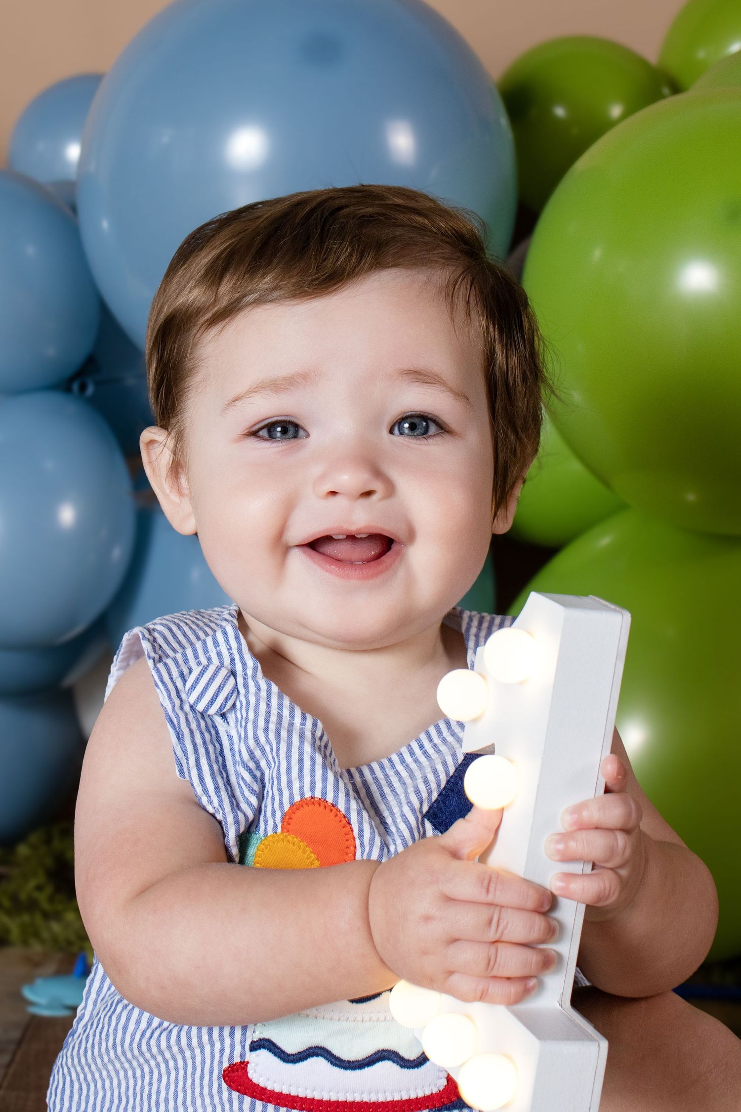 Smiling baby celebrating first birthday, holding number one, with blue and green balloons in background.