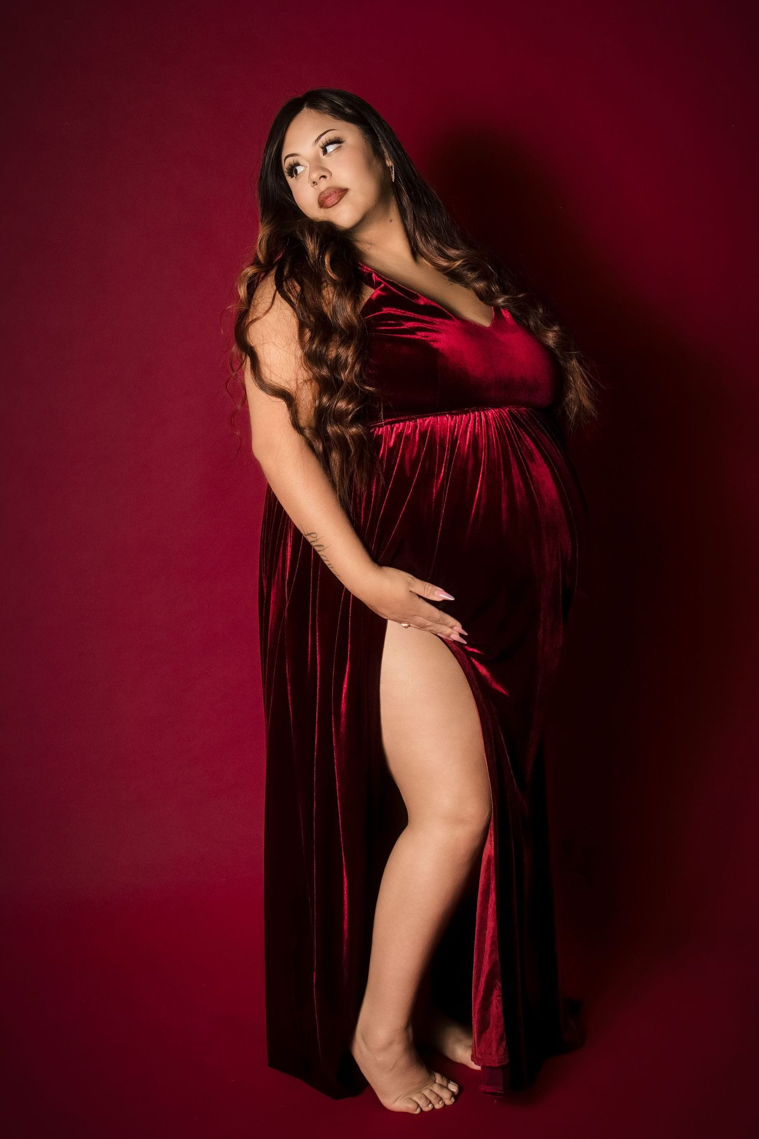 Pregnant woman in elegant red velvet gown with thigh slit posing against deep red background in maternity photo shoot.