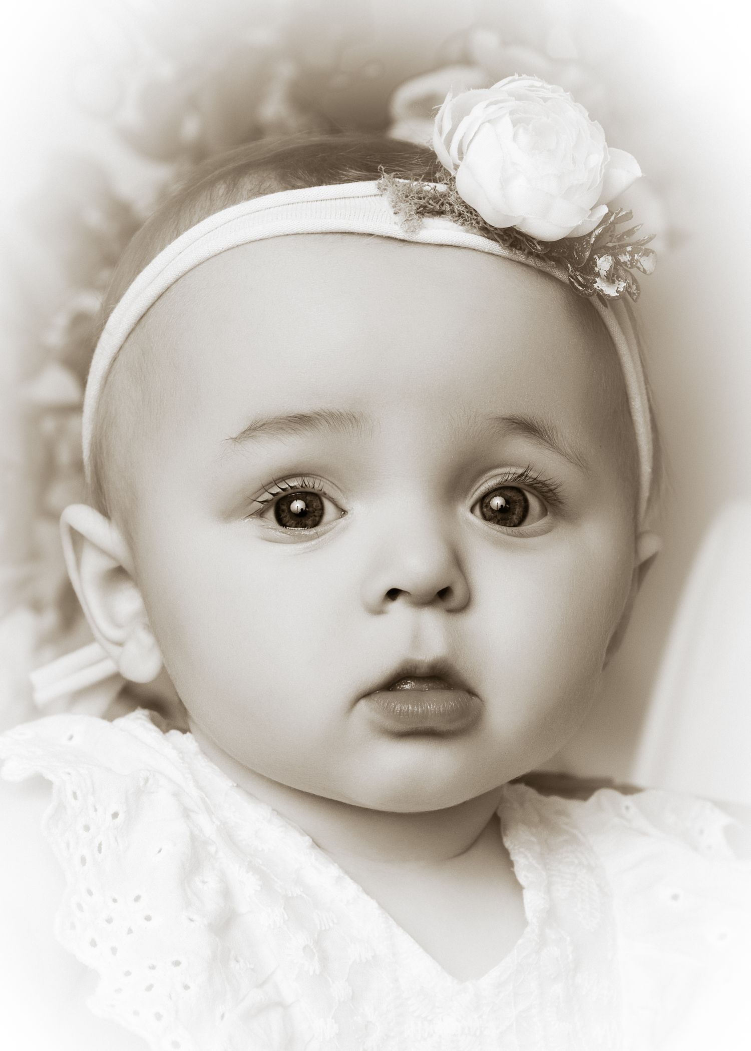 Sepia portrait of an adorable baby girl wearing a white floral headband and lace dress, with big expressive brown eyes.