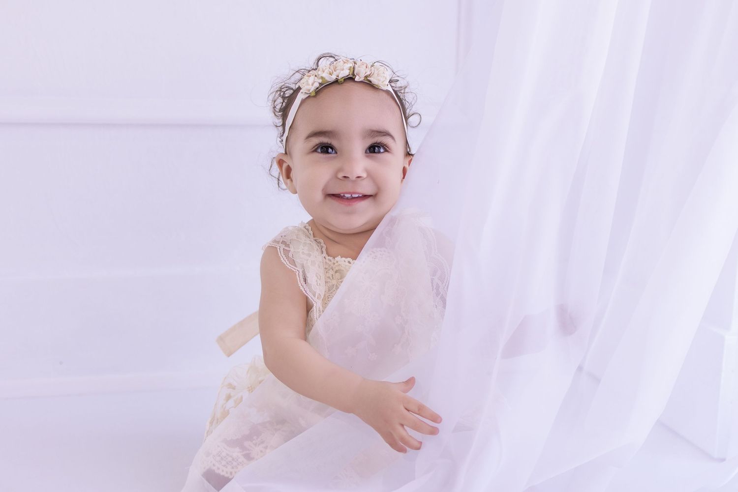 Smiling toddler girl in white lace dress and floral headband posing near sheer white curtains in bright studio.