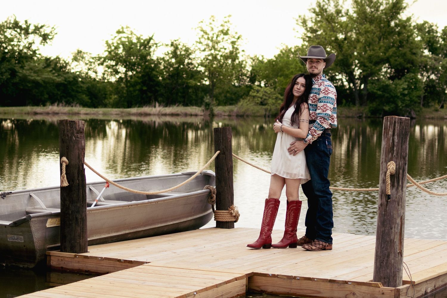Couple embracing on a wooden dock by a calm lake with a canoe, woman in red boots and white dress.