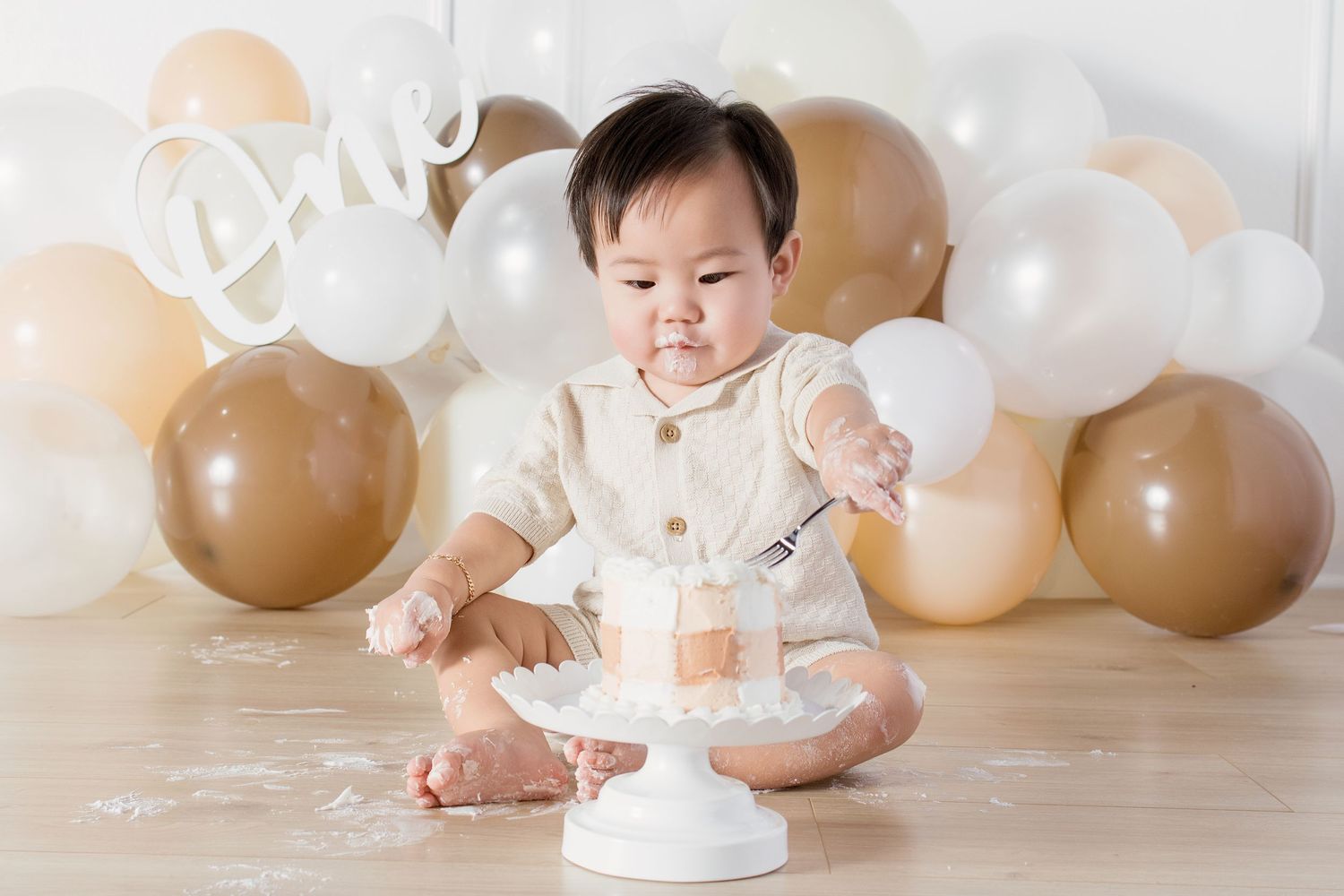 Baby in cream outfit smashes first birthday cake surrounded by gold and white balloons with One sign.
