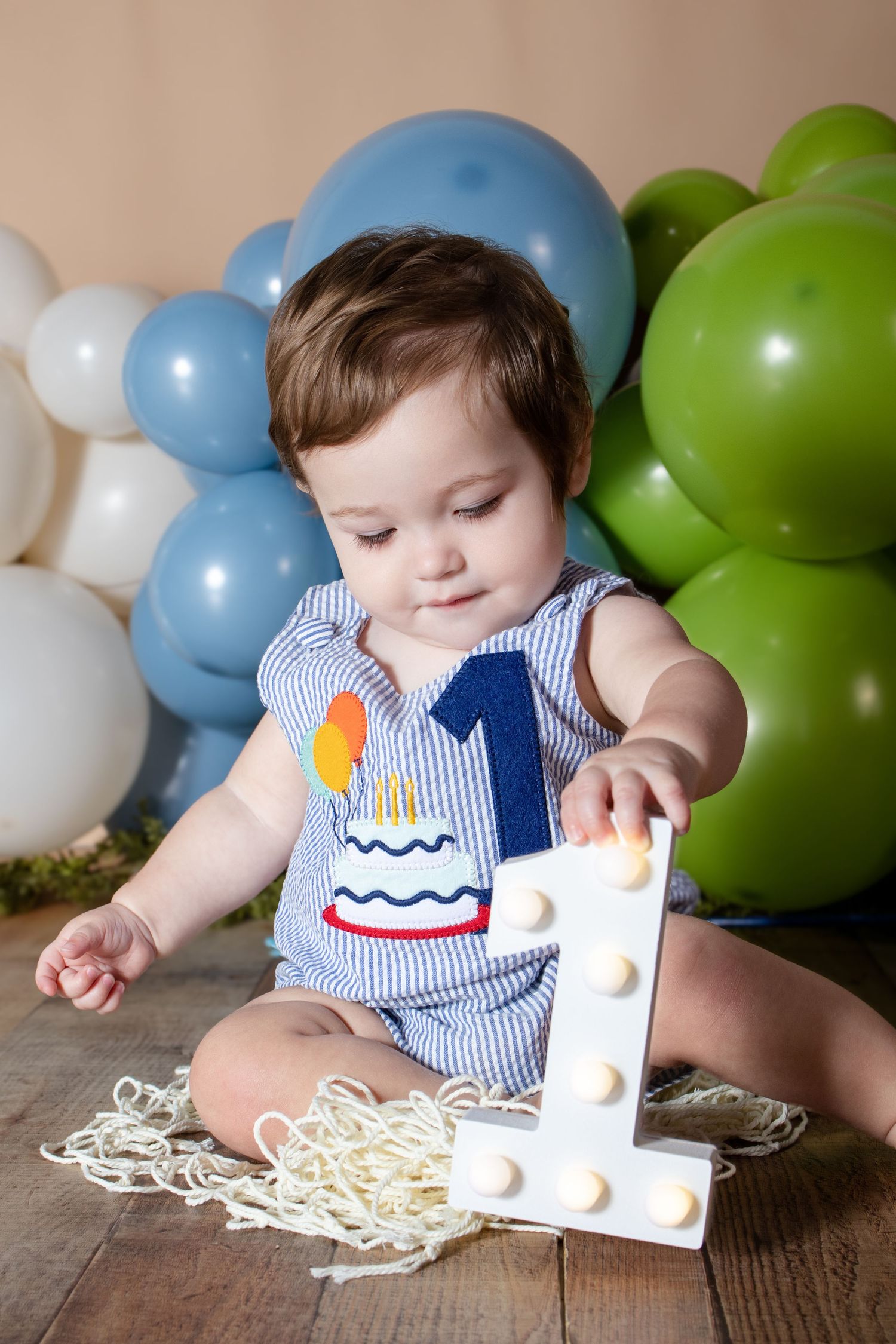 Baby boy in striped birthday romper with number 1 reaches for illuminated letter prop surrounded by blue and green balloons.