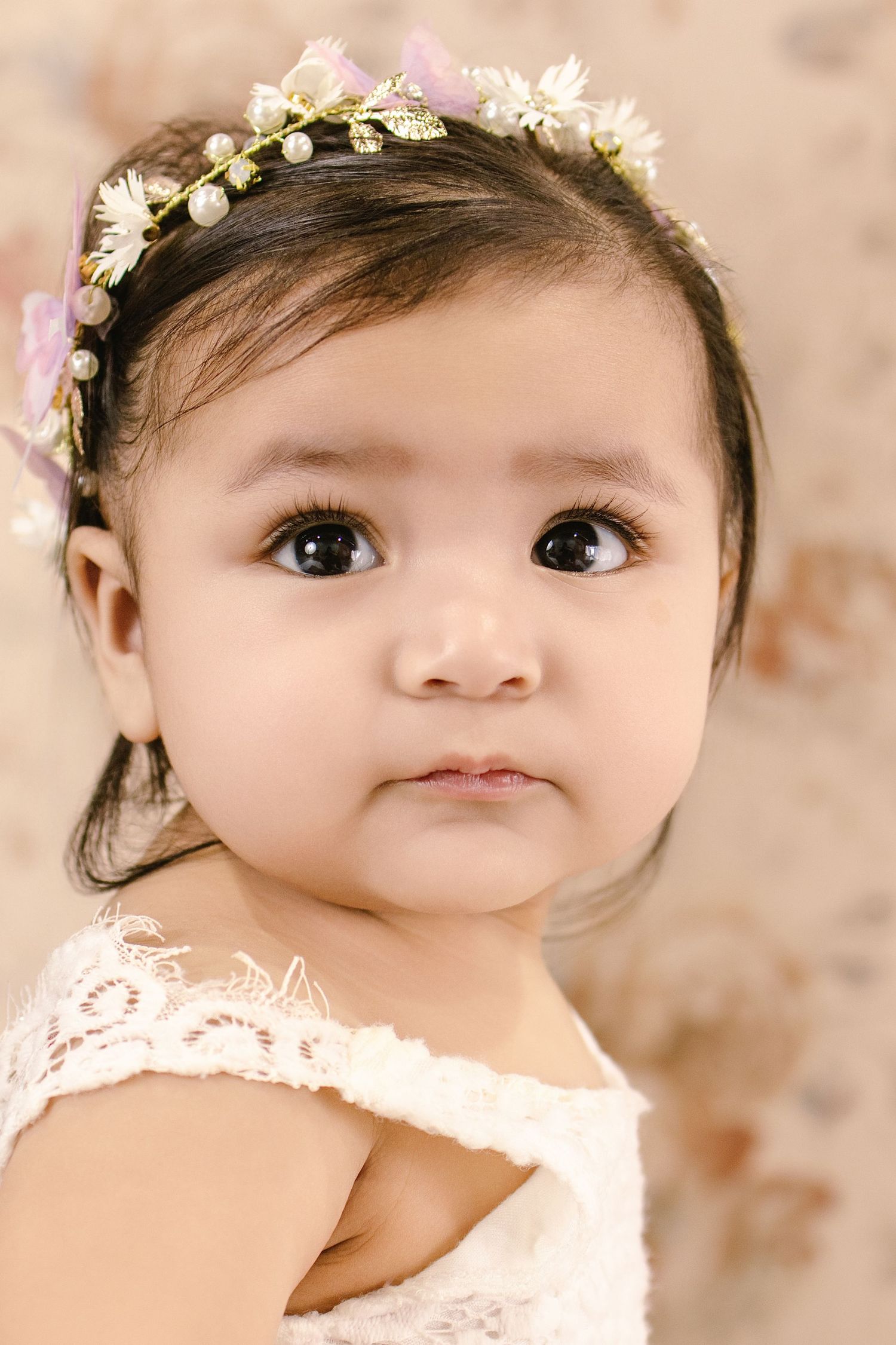 Adorable baby girl wearing a floral pearl headband and white lace dress, with big brown eyes and dark hair.