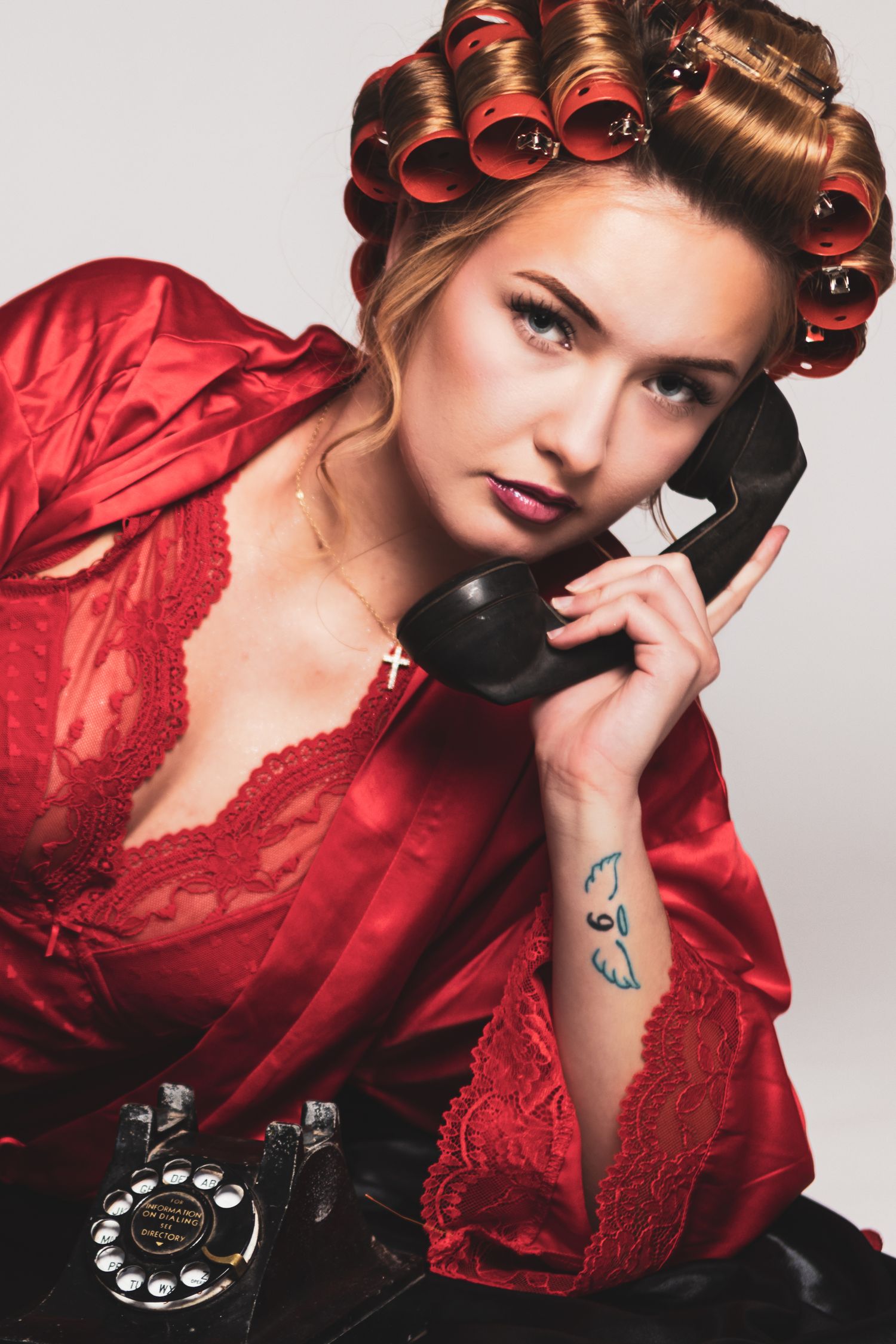 Woman in red satin robe with hair curlers talks on a vintage rotary phone, tattoo visible on wrist.
