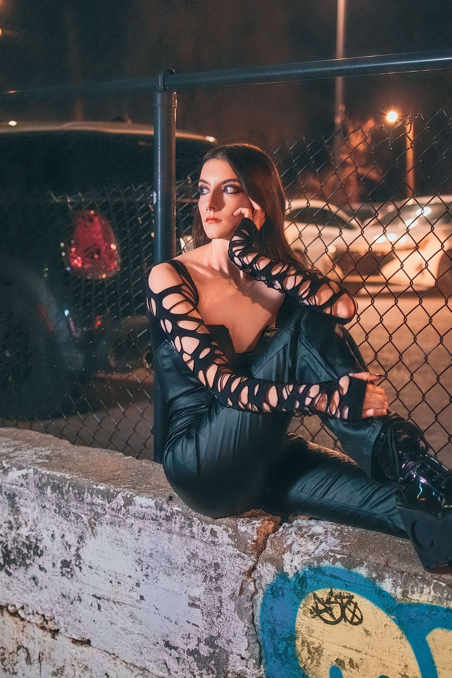 Woman in black lace-sleeve outfit sits against chain-link fence with city lights at night.