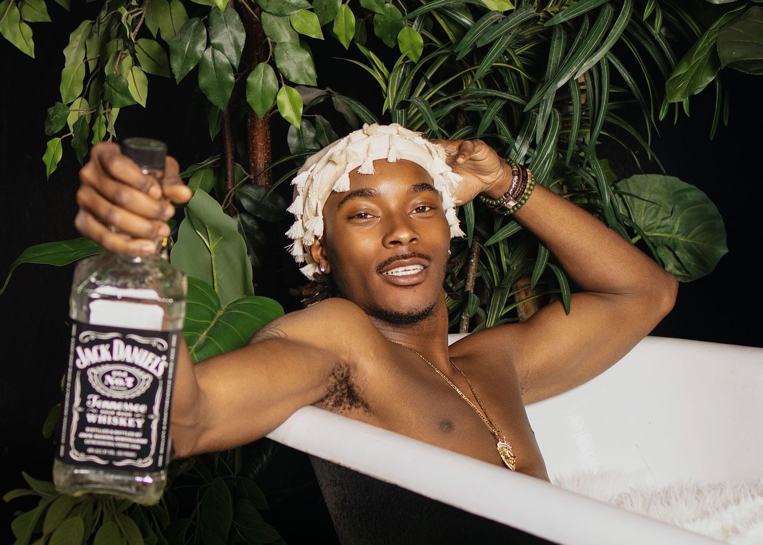 Shirtless man holding Jack Daniels whiskey bottle in a bathtub, wearing shower cap, surrounded by green plants.