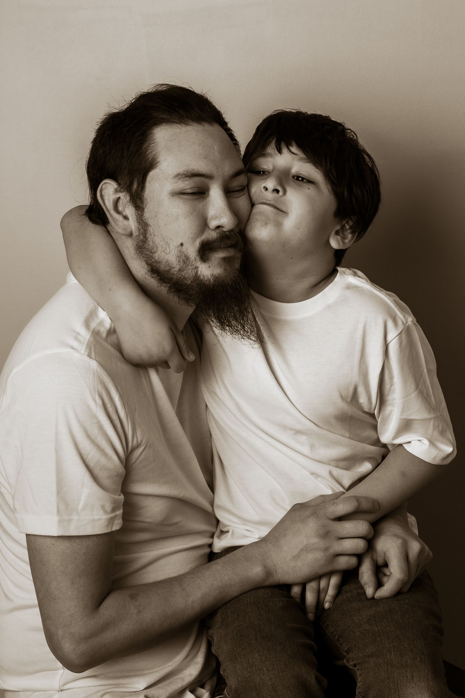 Sepia-toned portrait of a bearded father and young son sharing a tender embrace in white t-shirts.