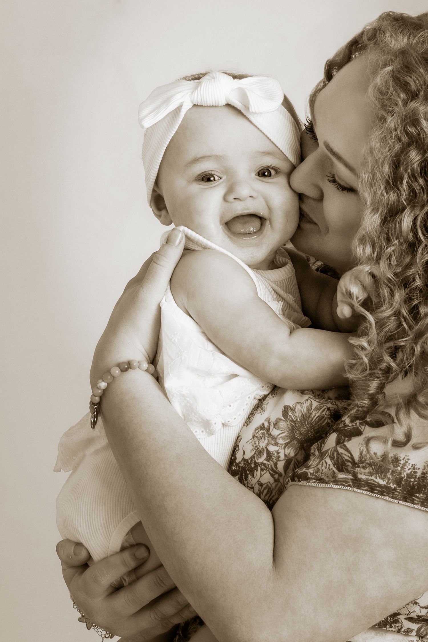 Sepia-toned photo of a laughing baby girl in white bow headband being kissed by her curly-haired mother.