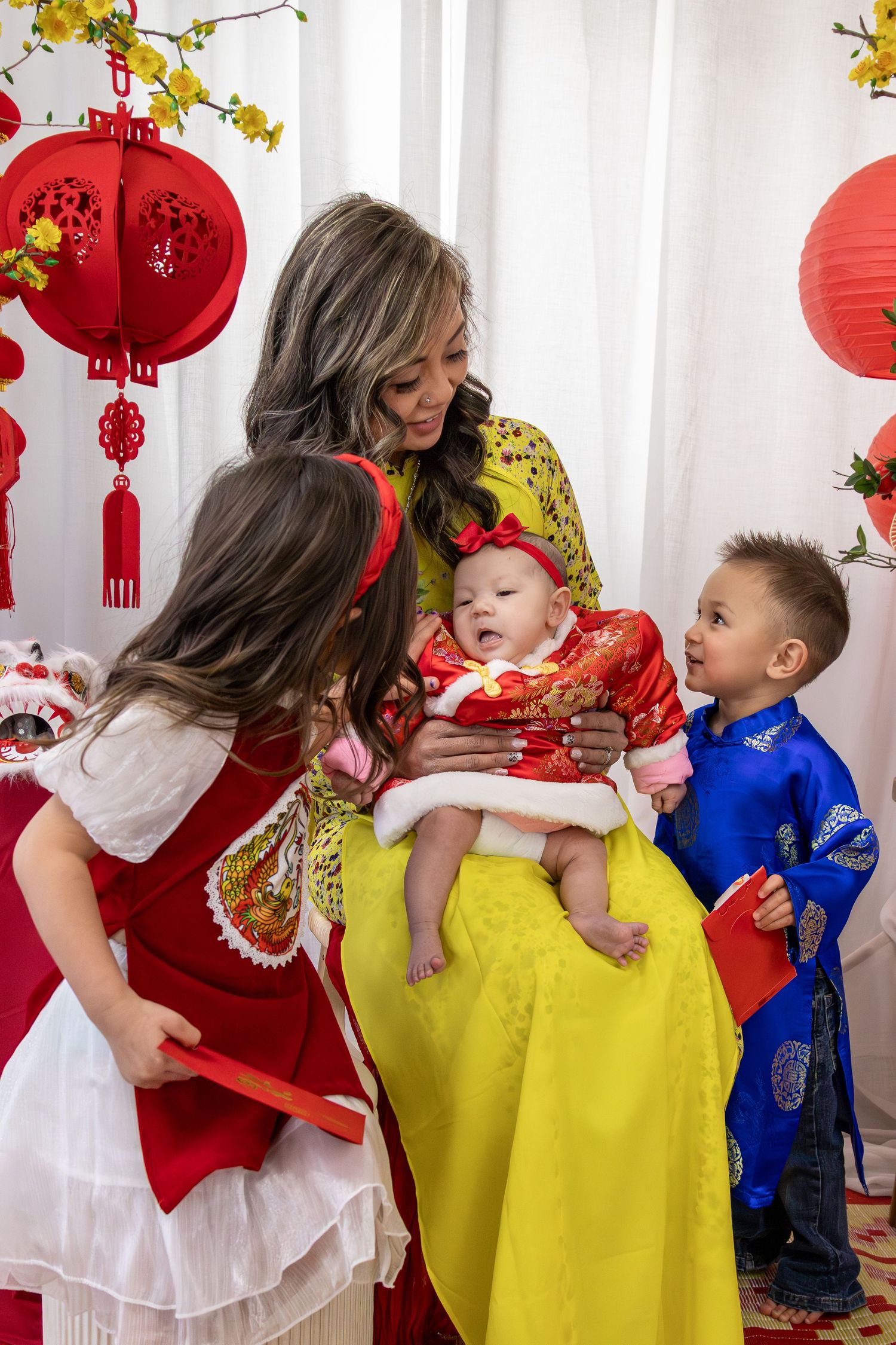 Family celebrating Lunar New Year in traditional Vietnamese ao dai outfits, with red lanterns and decorations in background.