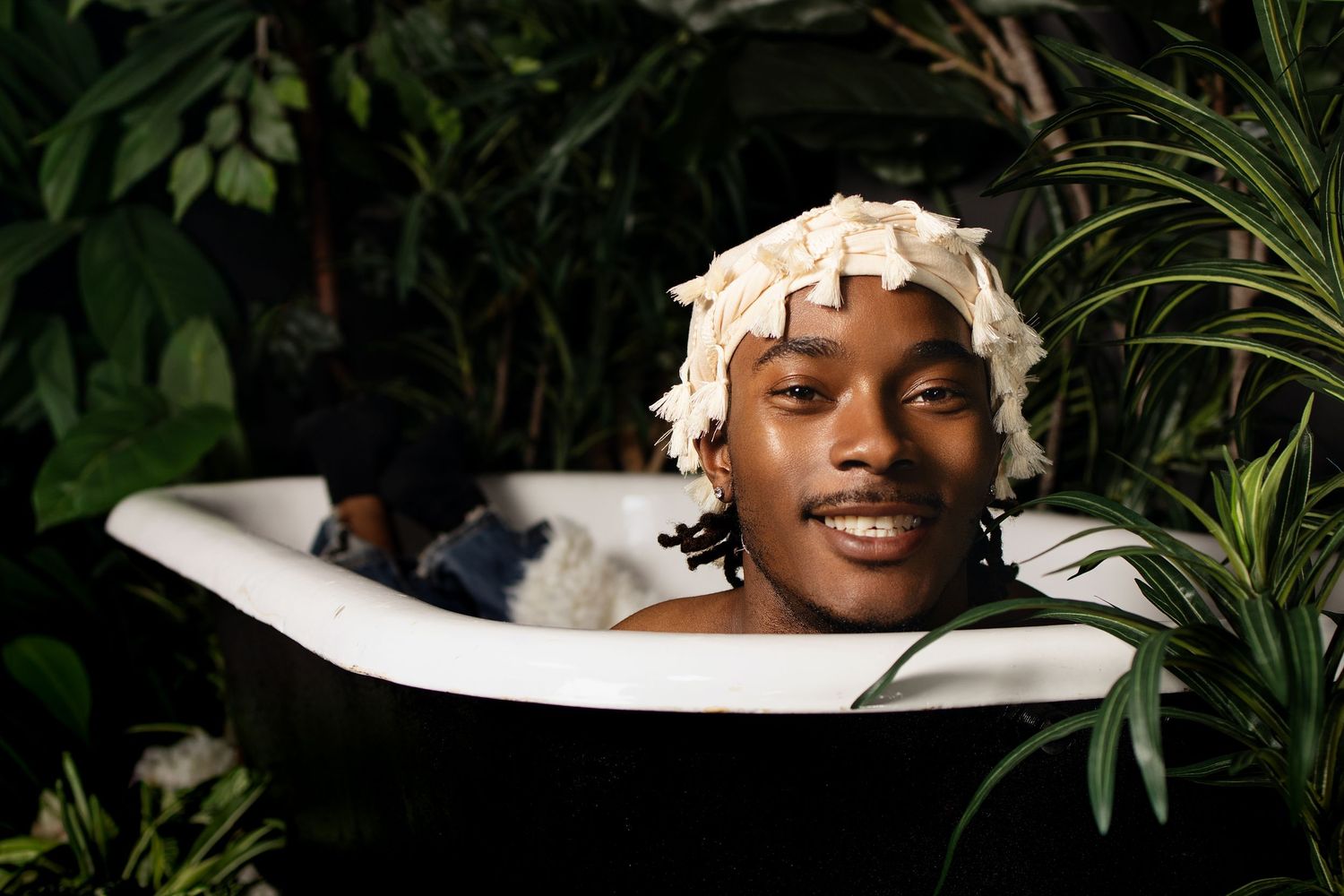 Young man smiling in a black bathtub wearing a shower cap, surrounded by lush green tropical plants.
