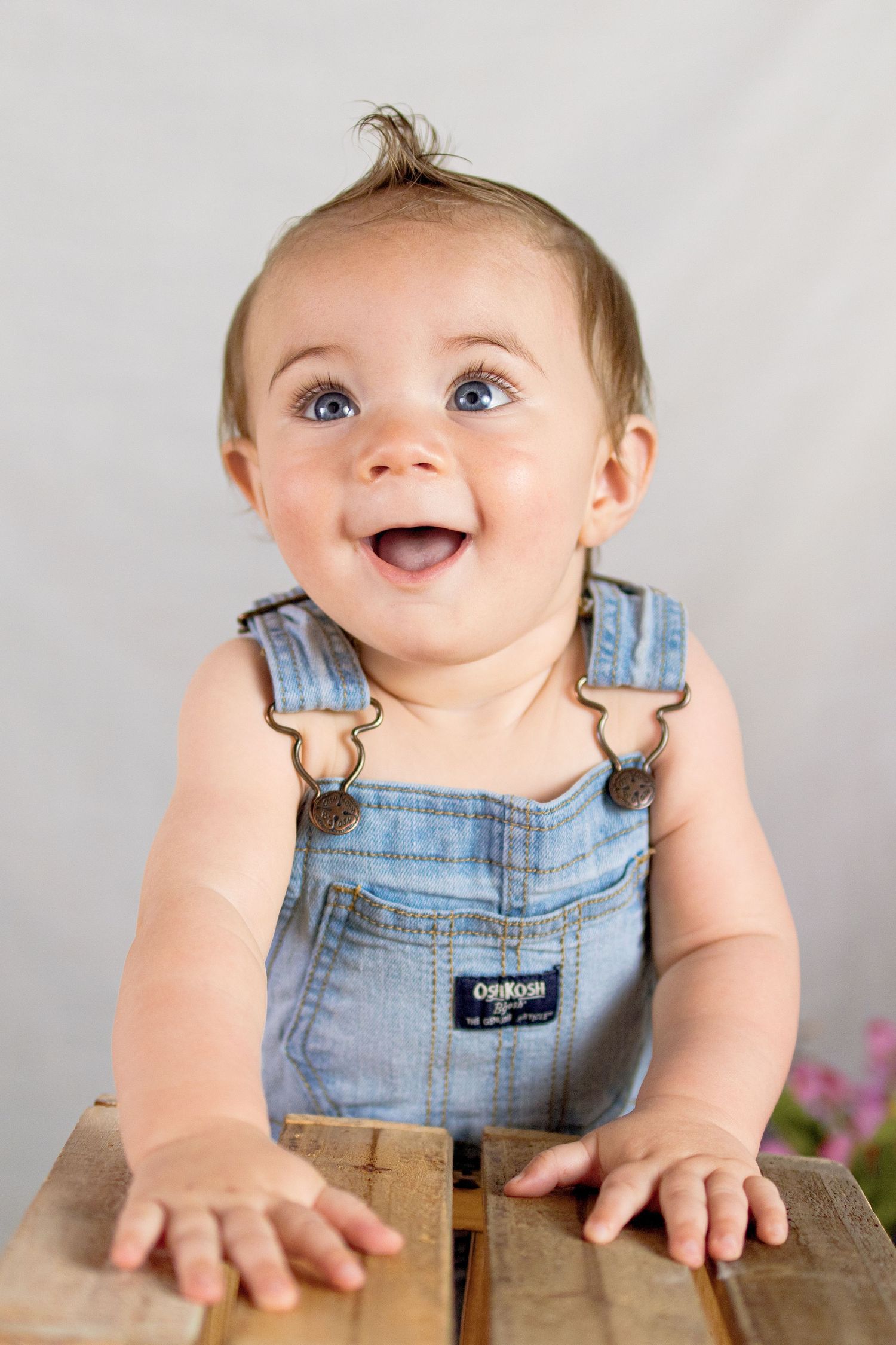 Happy baby with blue eyes wearing denim OshKosh overalls, laughing while leaning on wooden crate.