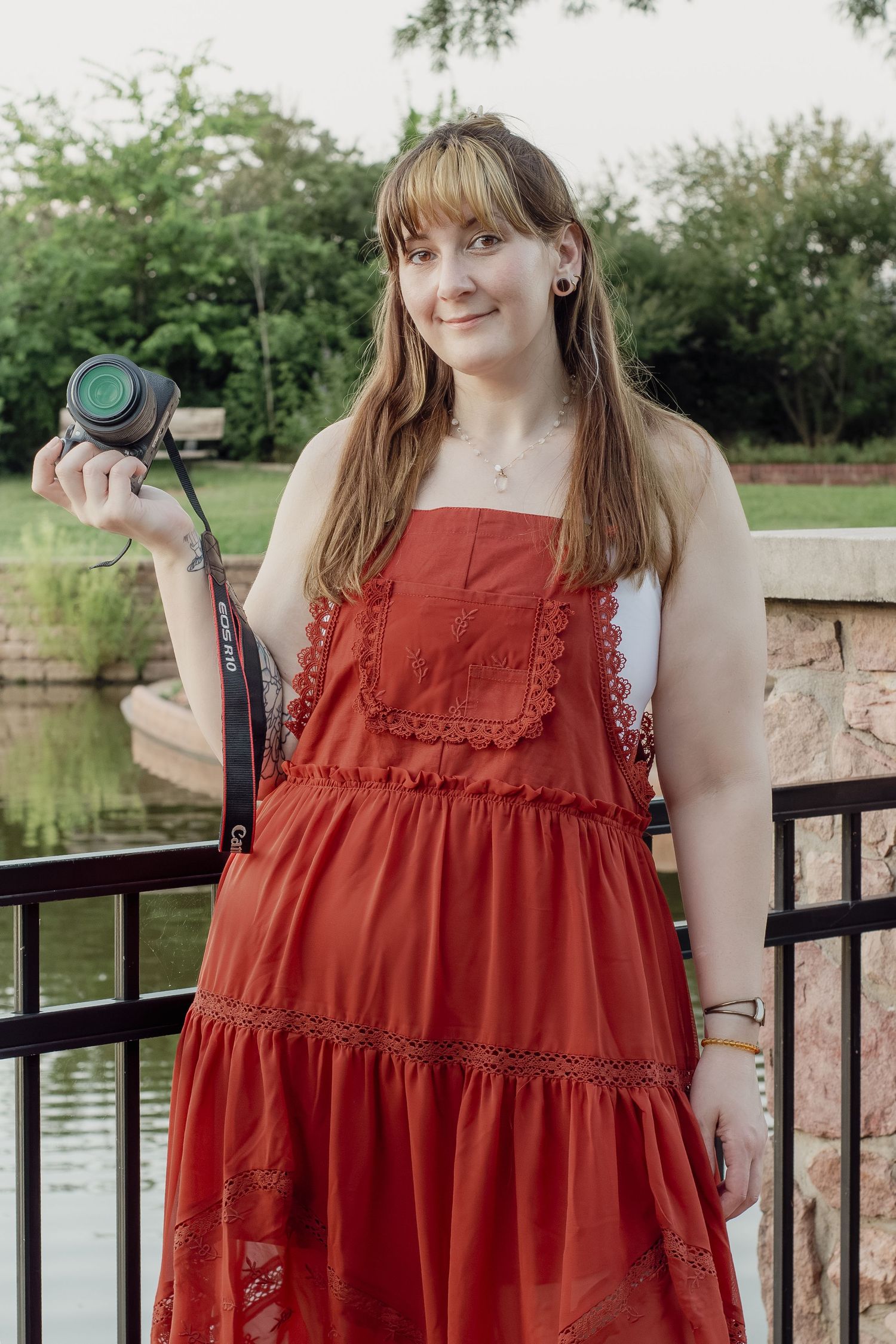 Young woman in red dress holding a camera outdoors near a pond with green trees in background.