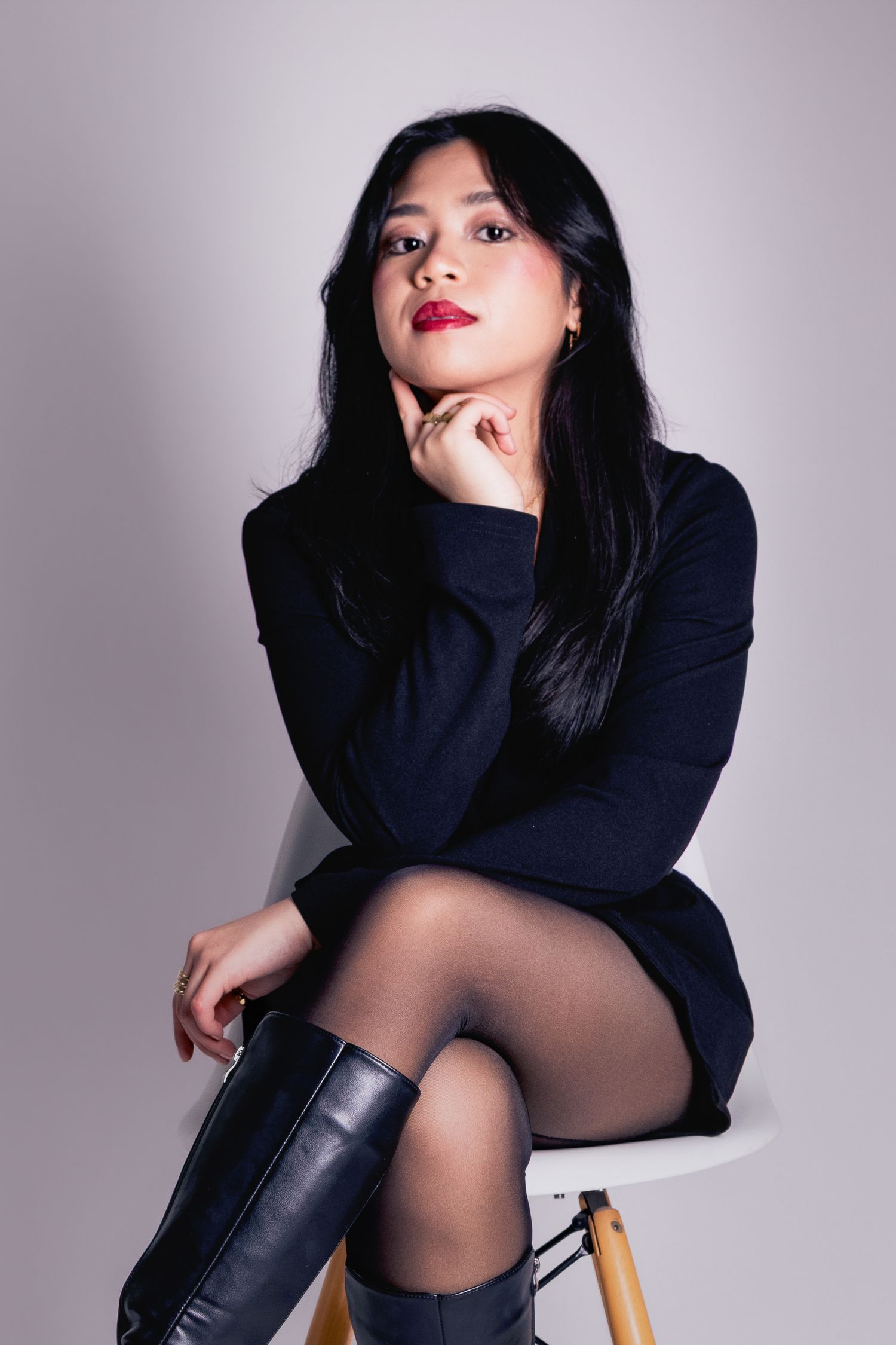 Woman in black dress and knee-high boots seated on chair, resting chin on hand, red lips, studio portrait.