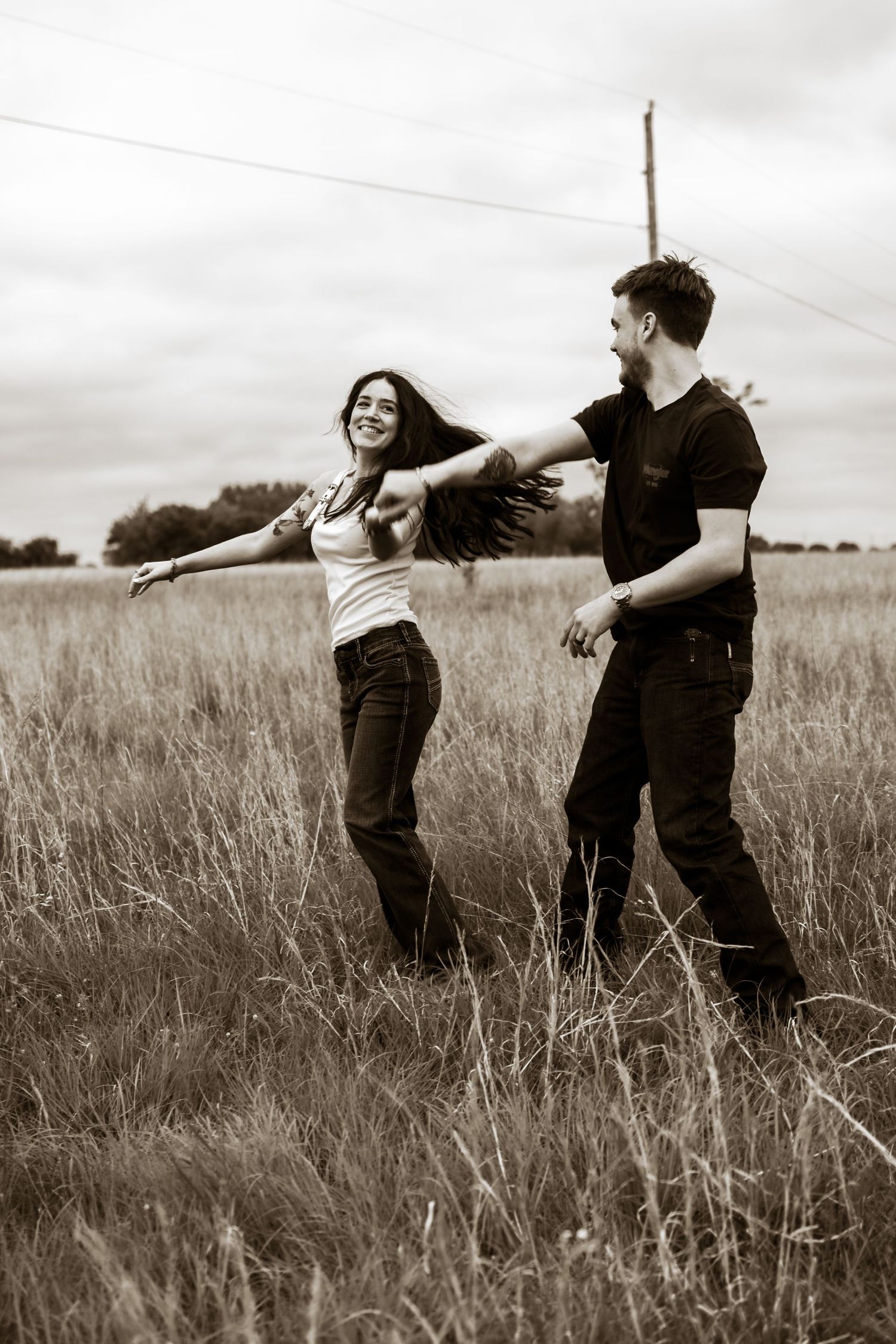 Couple dancing joyfully in a golden field, woman spinning with long hair flowing, sepia-toned romantic outdoor photo.