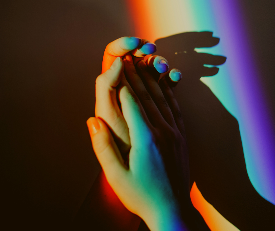 Two hands with colorful nail polish touching under vibrant rainbow light on a dark background.