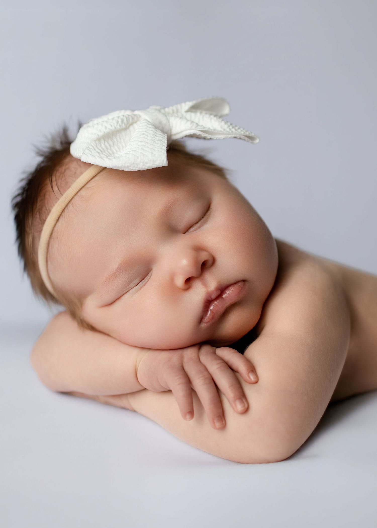 Sleeping newborn baby girl wearing a white bow headband, resting peacefully on her hands against a soft gray background.