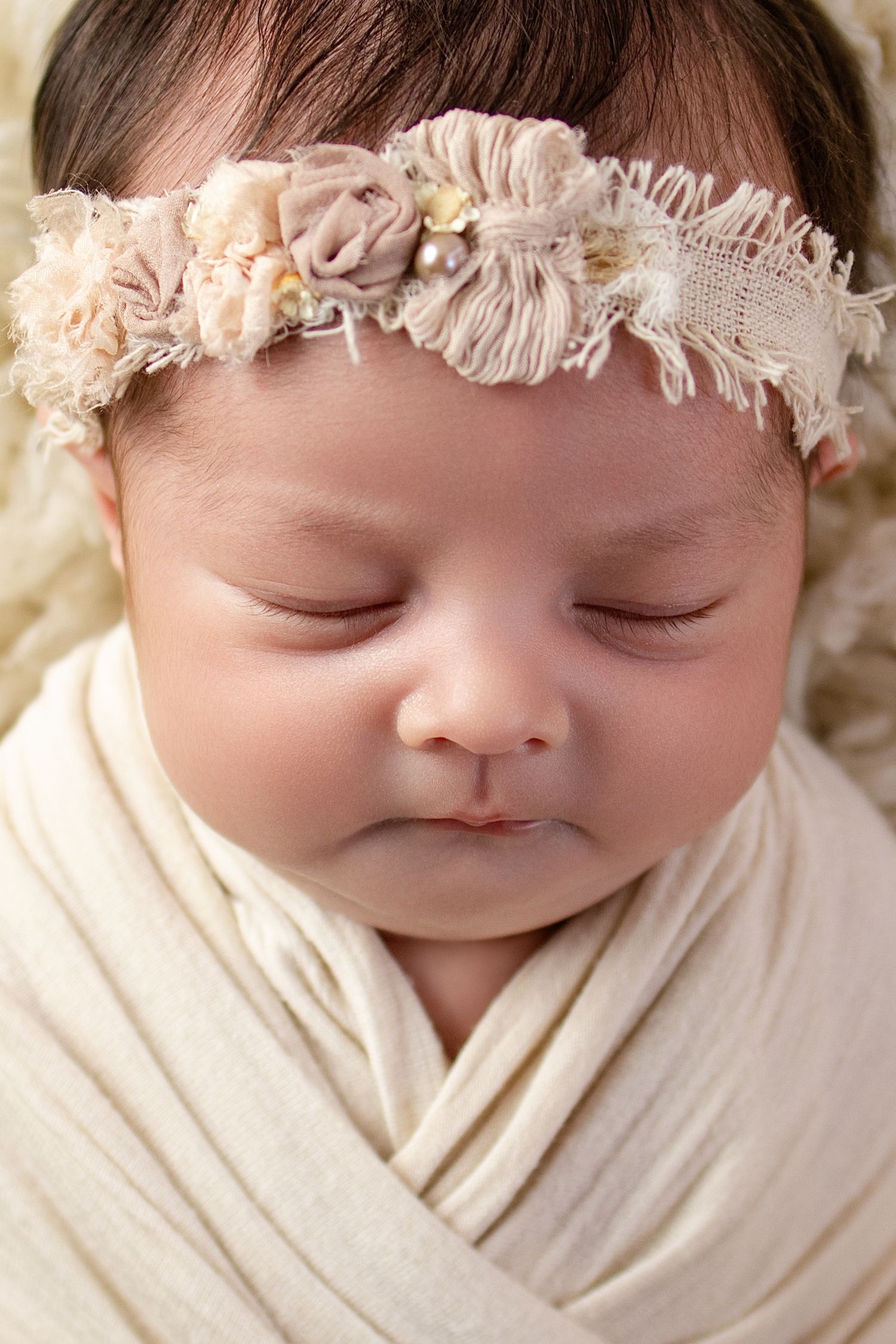 Sleeping newborn baby girl swaddled in cream wrap wearing a delicate floral headband, peaceful close-up portrait.