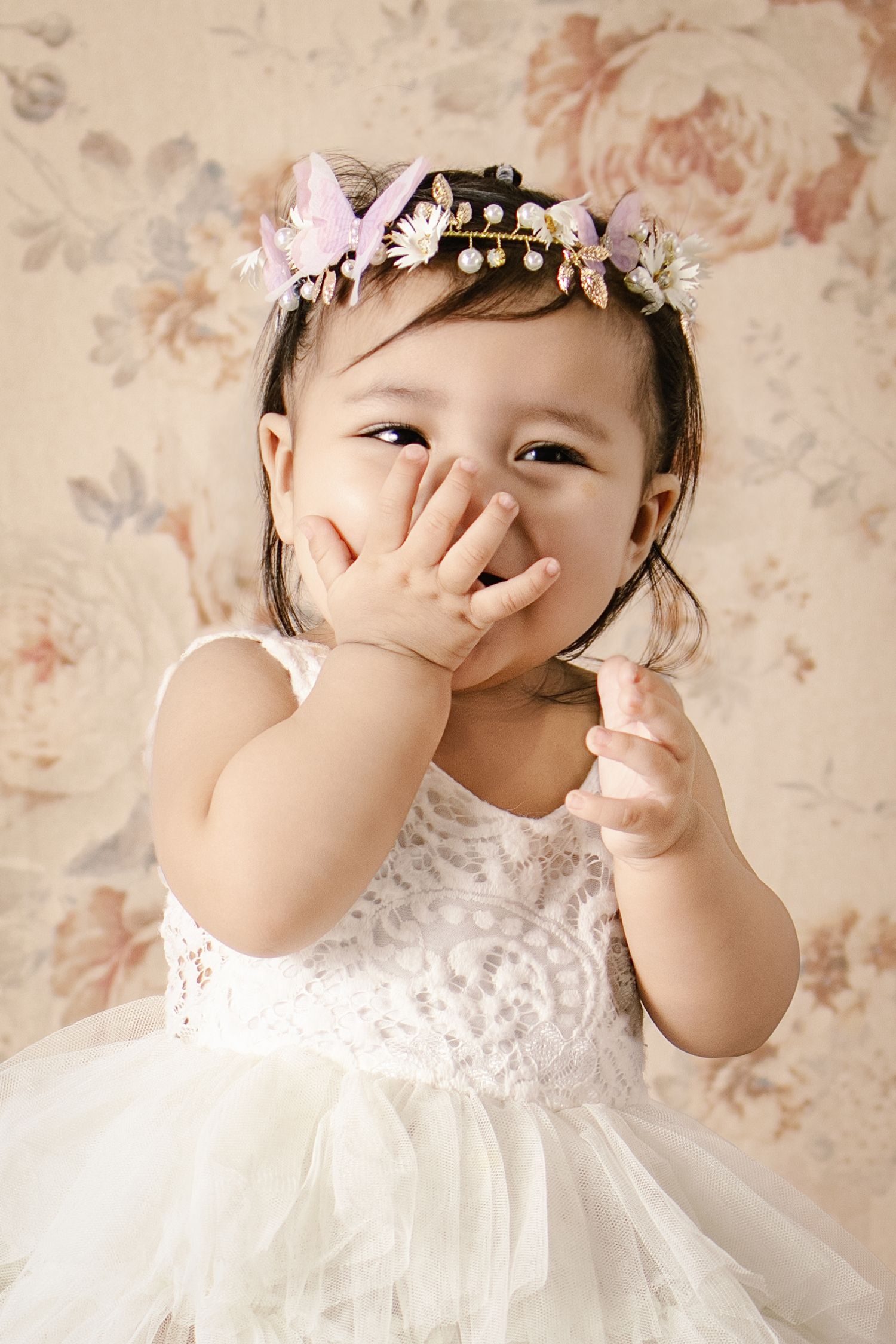 Adorable baby girl in white lace dress and floral crown, giggling with hand over mouth against floral backdrop.