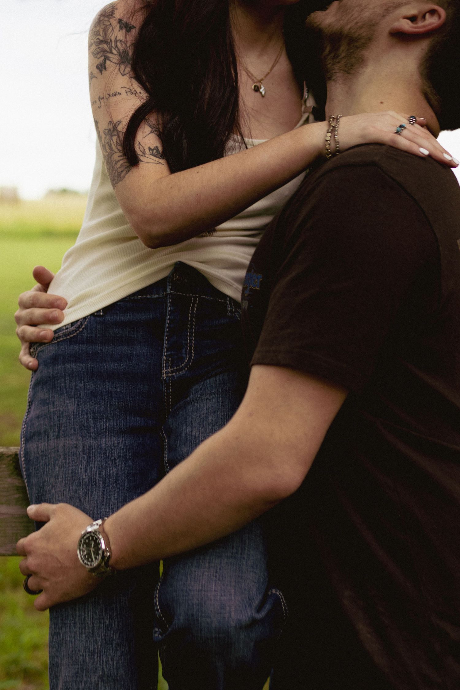 A tattooed couple embracing outdoors, woman in white top and jeans with arm around mans shoulders in black shirt.