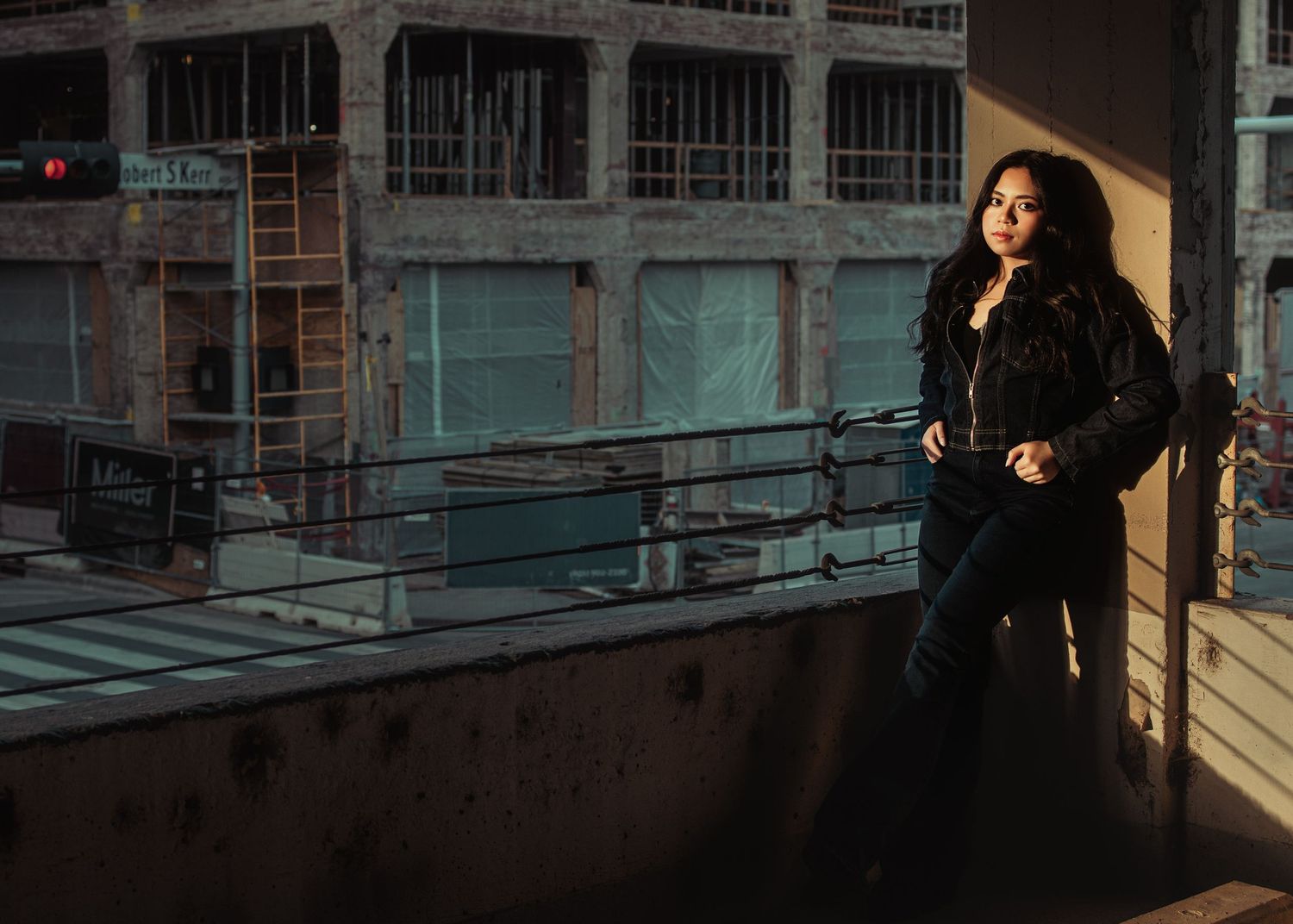 Woman in black jacket poses on urban rooftop terrace with industrial building backdrop at golden hour.