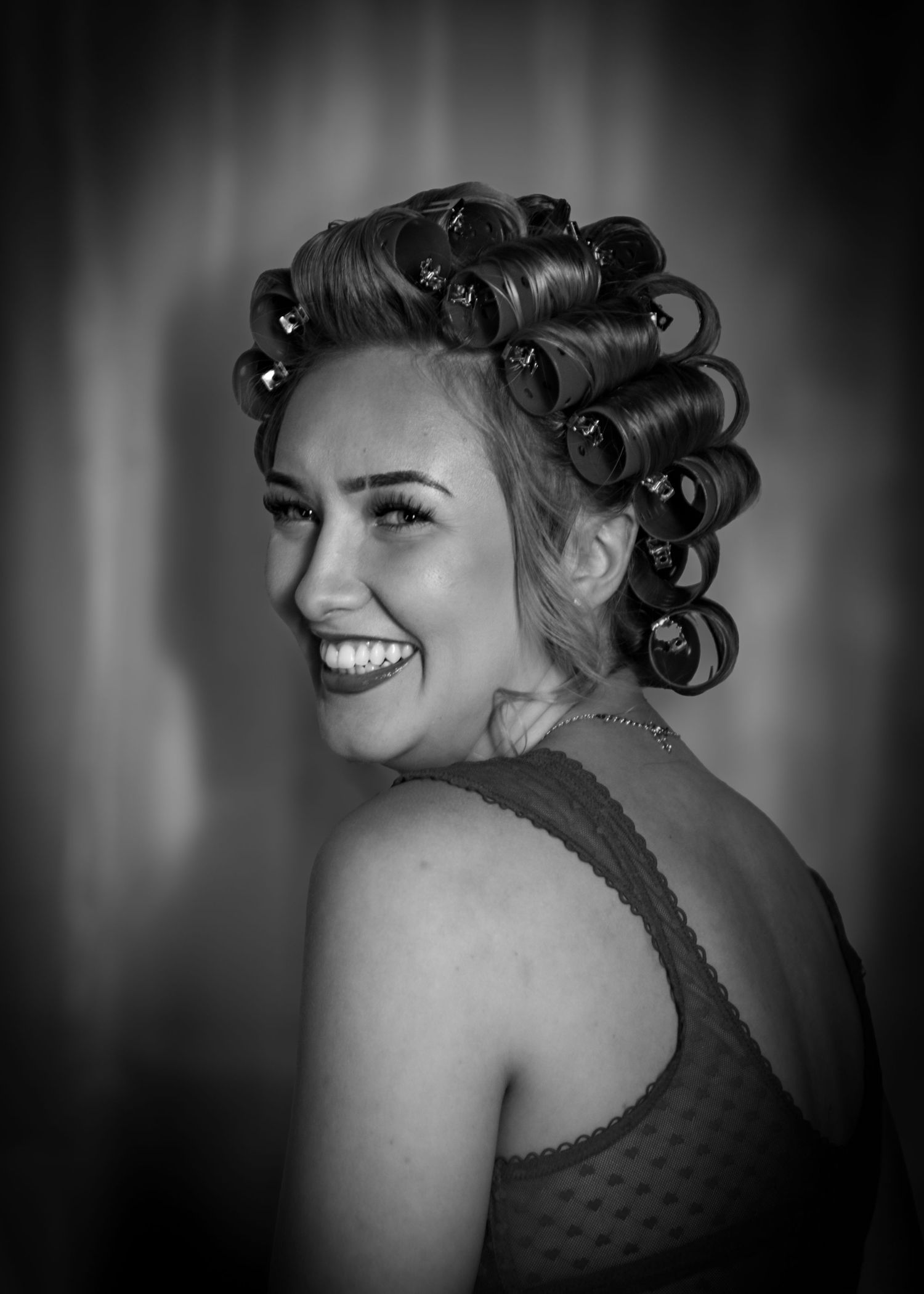 Black and white portrait of a smiling woman with hair rollers, wearing a lace top, looking over her shoulder.