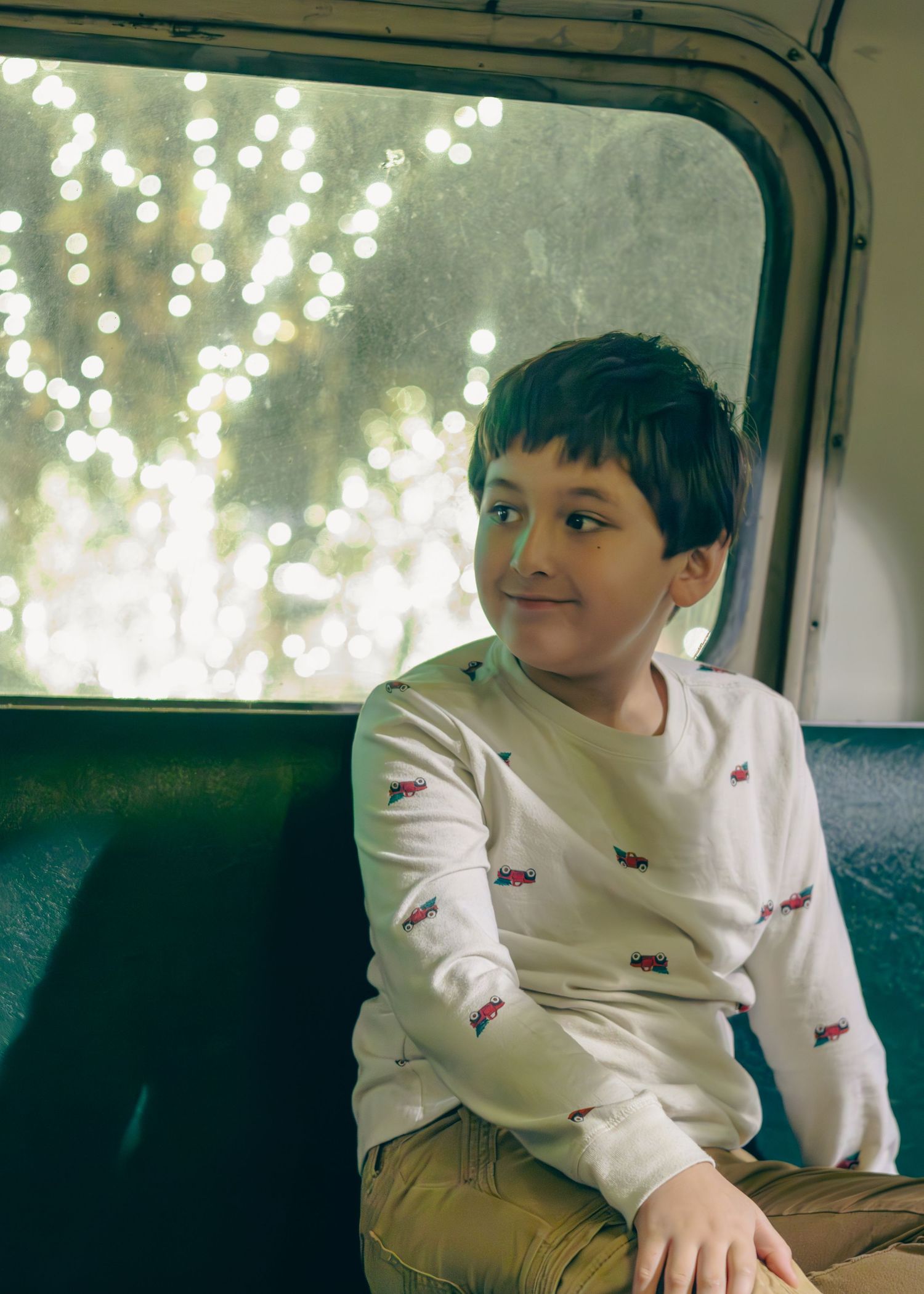 A young boy in a white long-sleeve shirt sits smiling on a train seat, gazing out a bokeh-lit window.