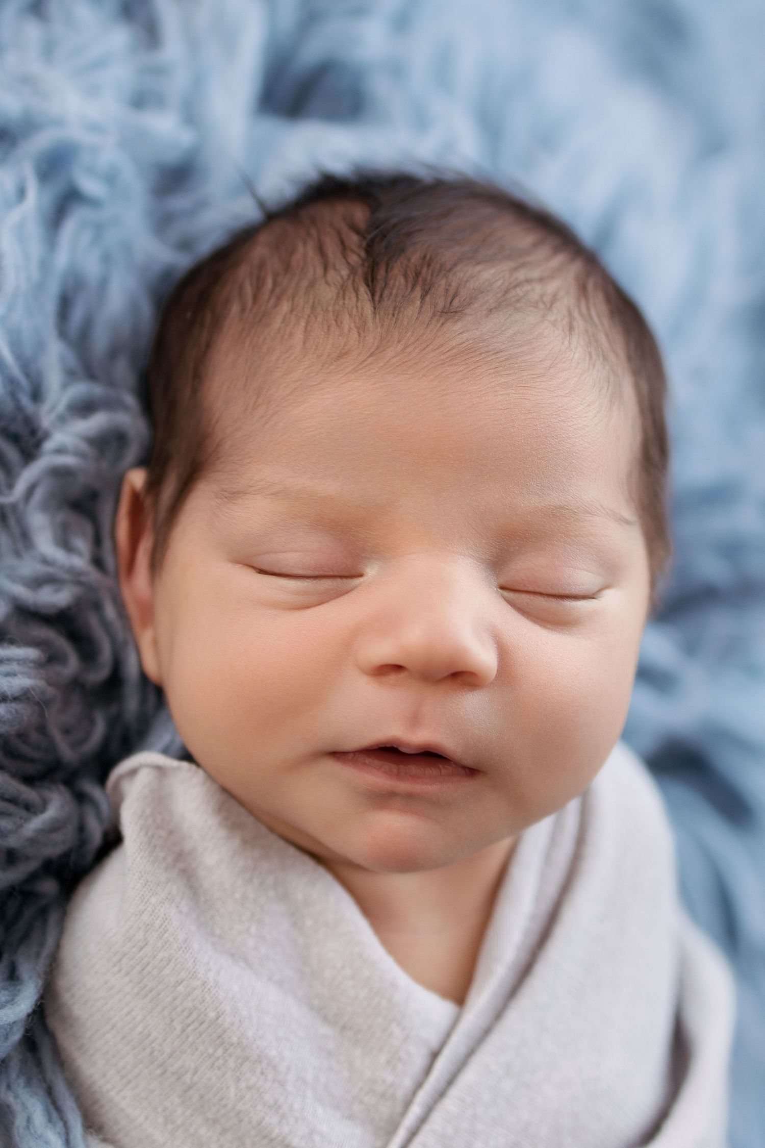 Peaceful newborn baby sleeping swaddled in gray wrap on soft blue fluffy blanket, close-up portrait.