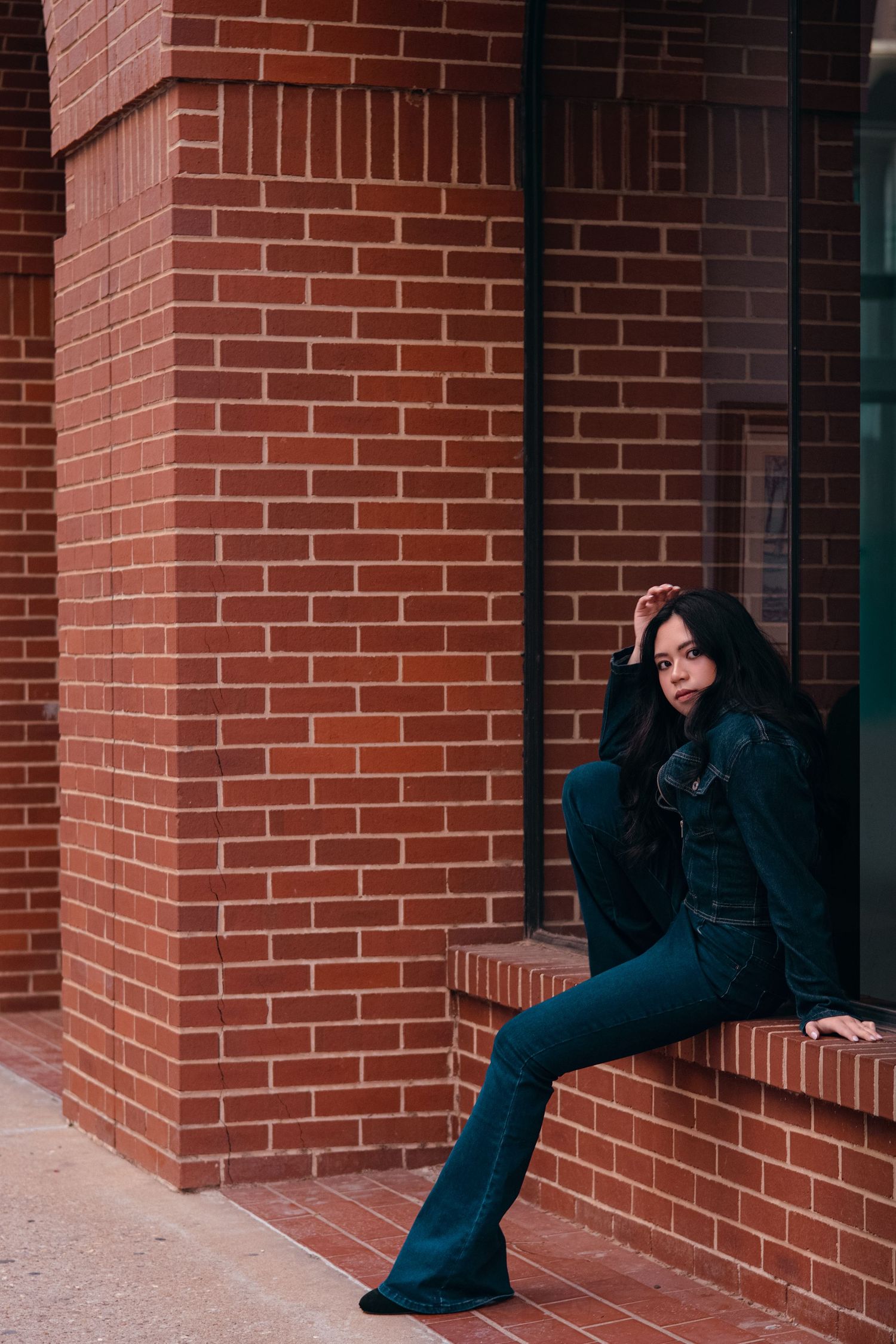 Young woman in dark denim outfit sits on brick building ledge, leaning against wall with legs extended.