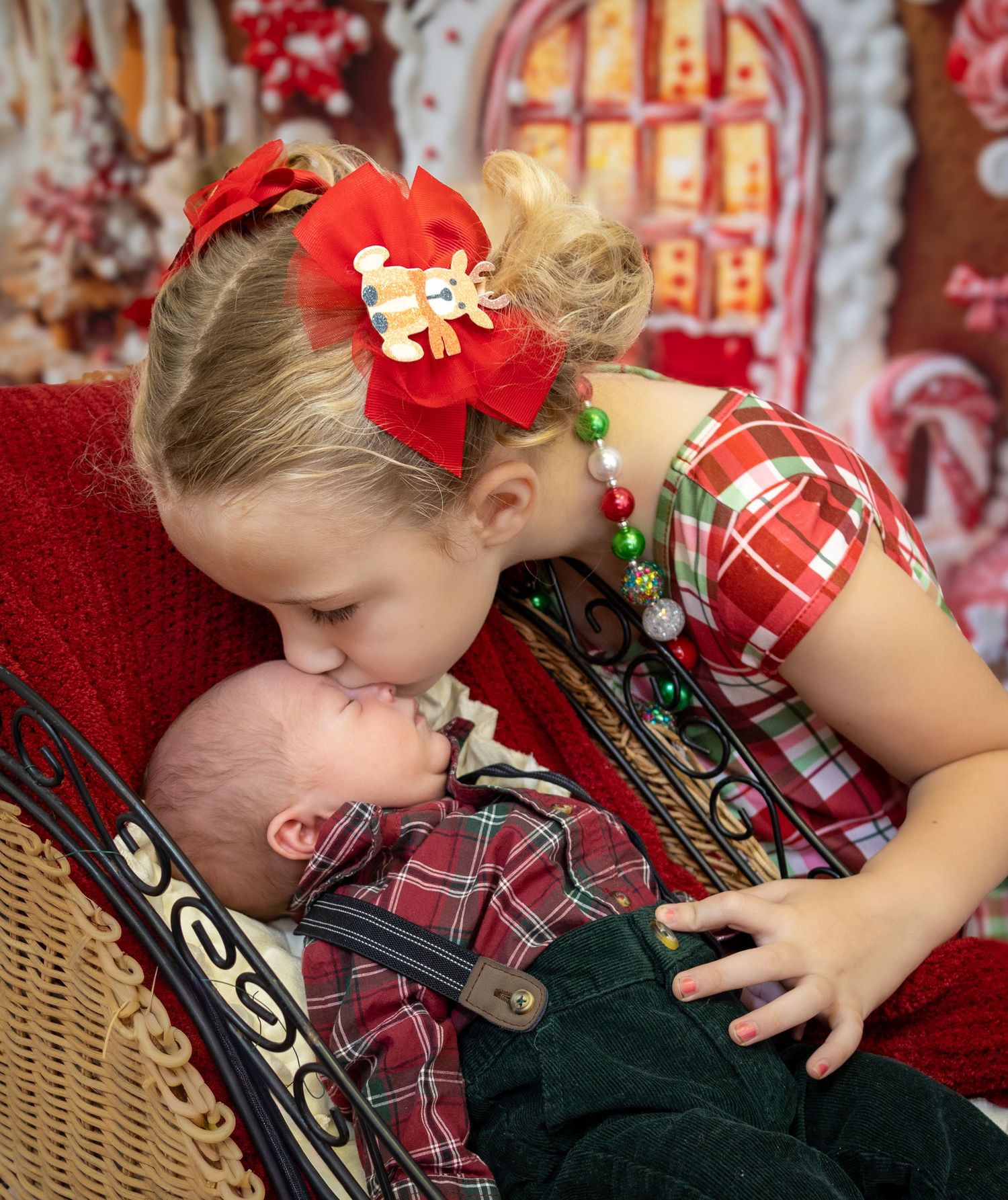 Girl in plaid Christmas dress kisses newborn baby in matching outfit on festive holiday backdrop.