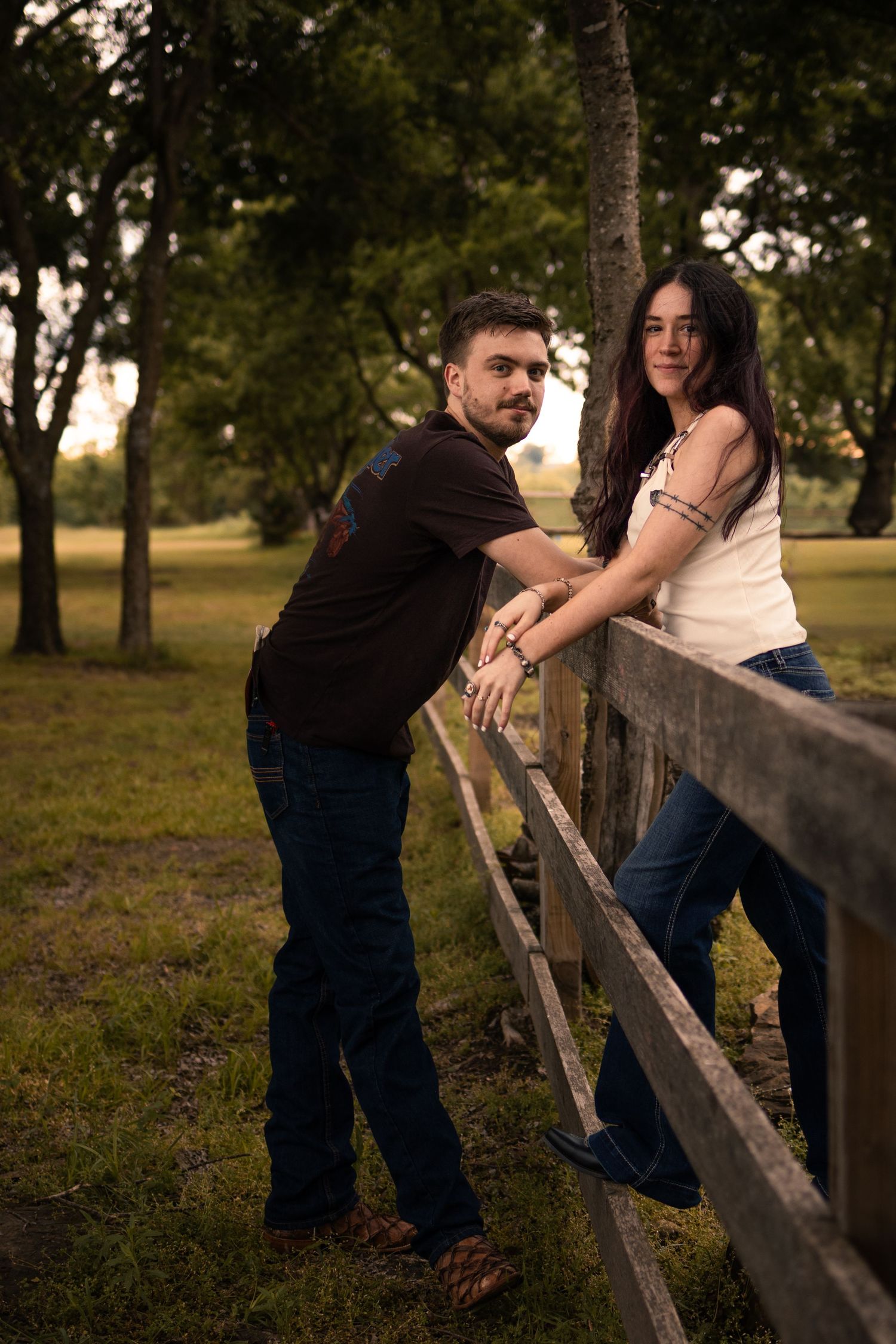 A young couple leans against a rustic wooden fence in a lush green park, posing together during golden hour.
