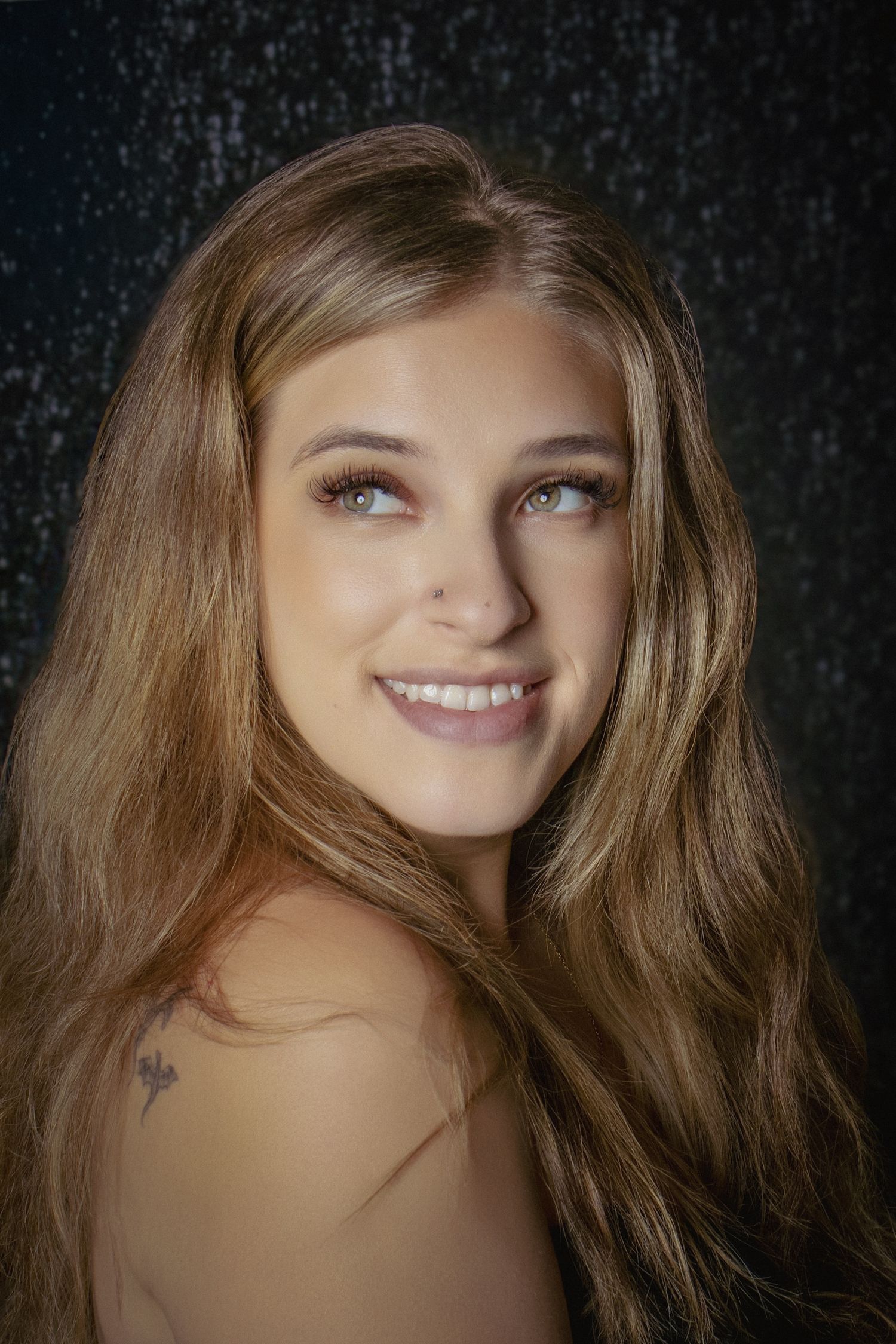 Smiling young woman with long blonde hair, blue eyes, and a shoulder tattoo poses against a dark glittery background.