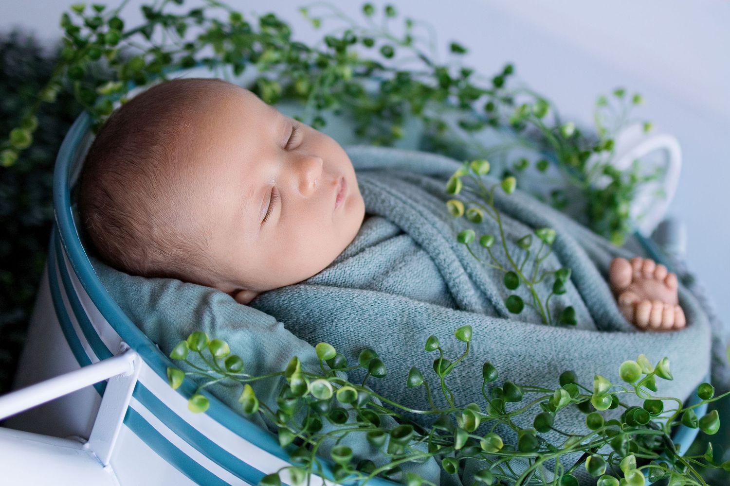 Sleeping newborn baby swaddled in a soft blue wrap, nestled in a basket surrounded by fresh green leaves.