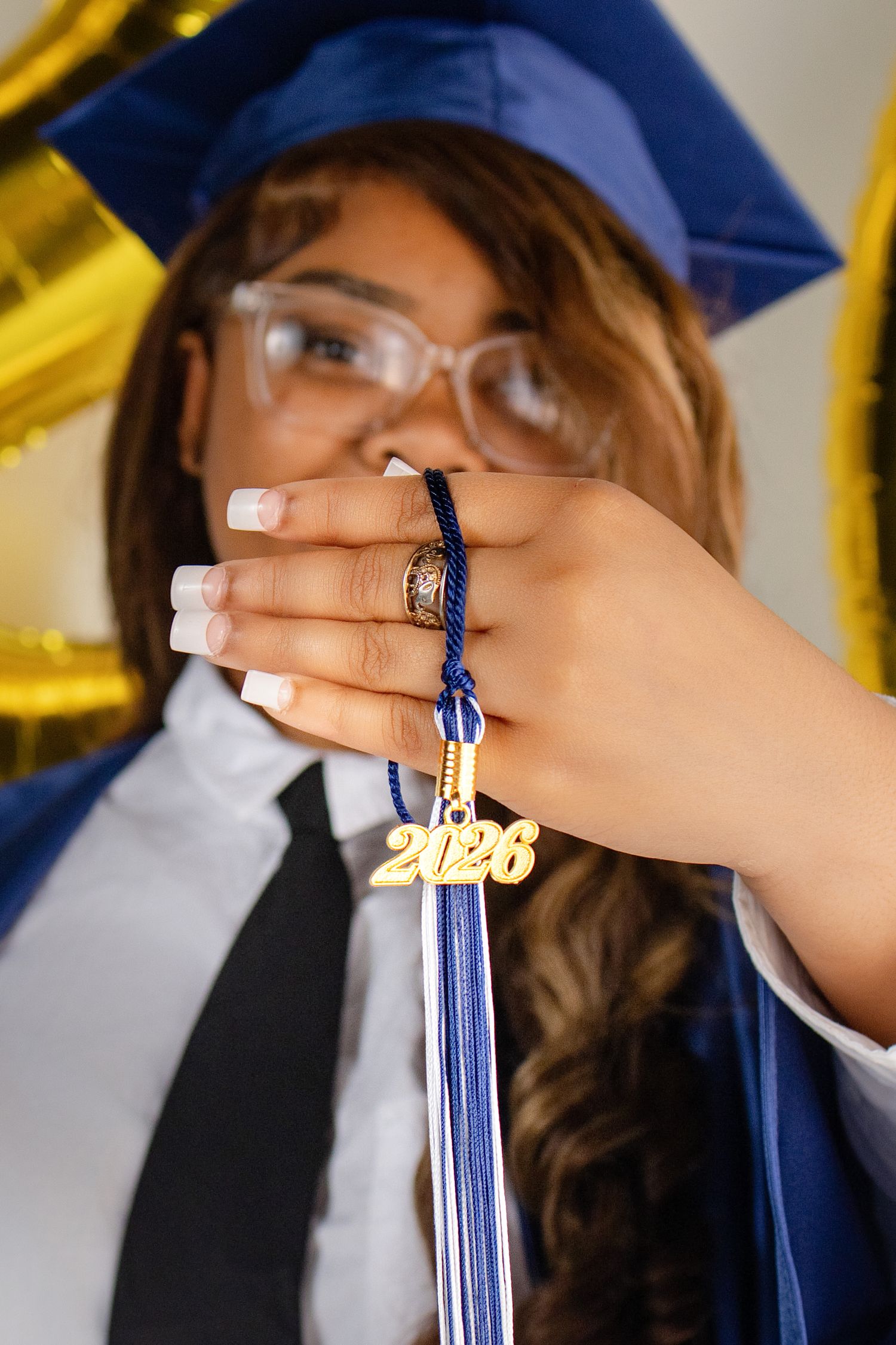 Graduate in blue cap and gown holds 2026 tassel, wearing glasses and white manicure, with gold balloons in background.