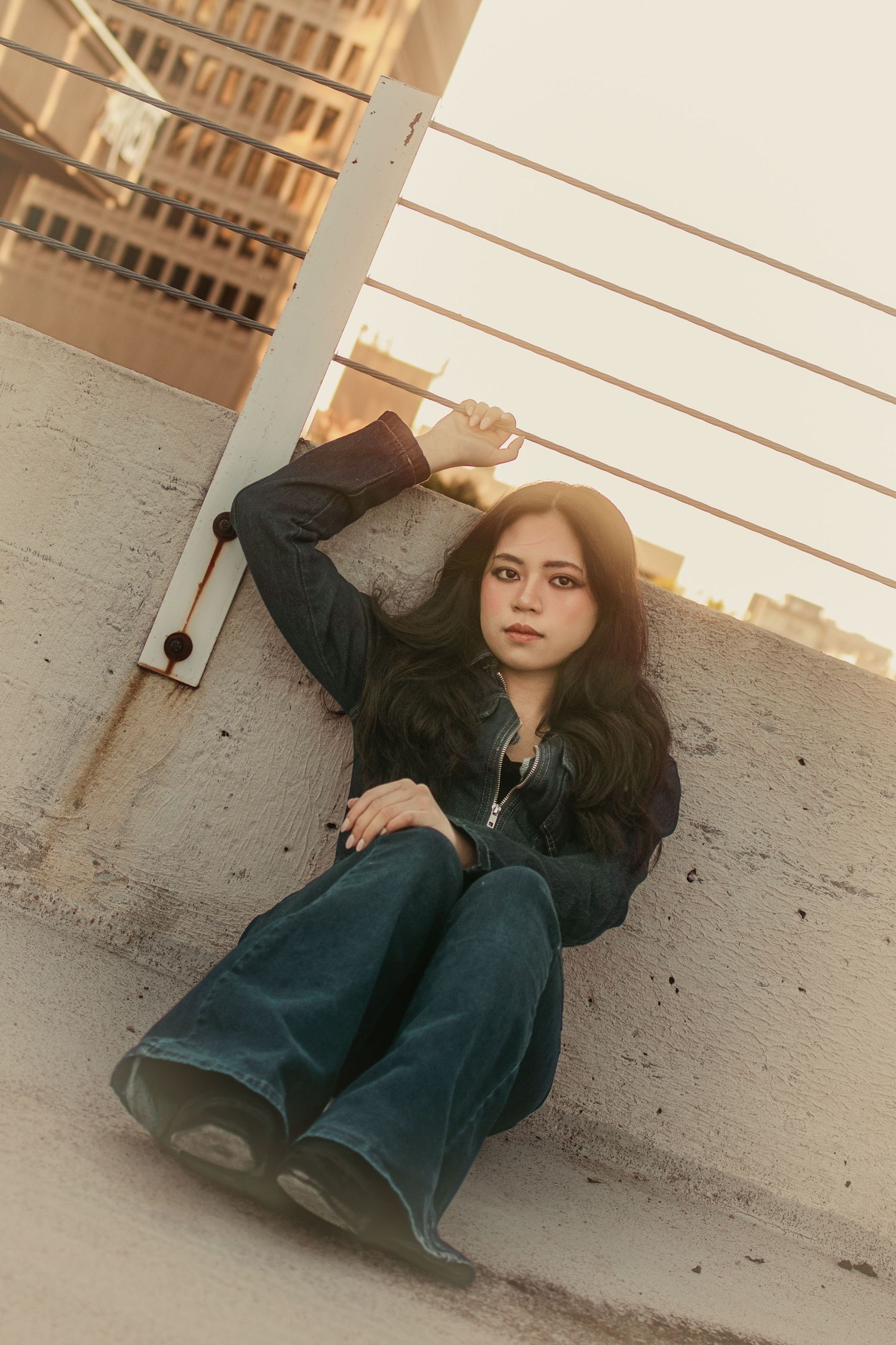 Woman in black jacket and wide-leg jeans sits against concrete wall.